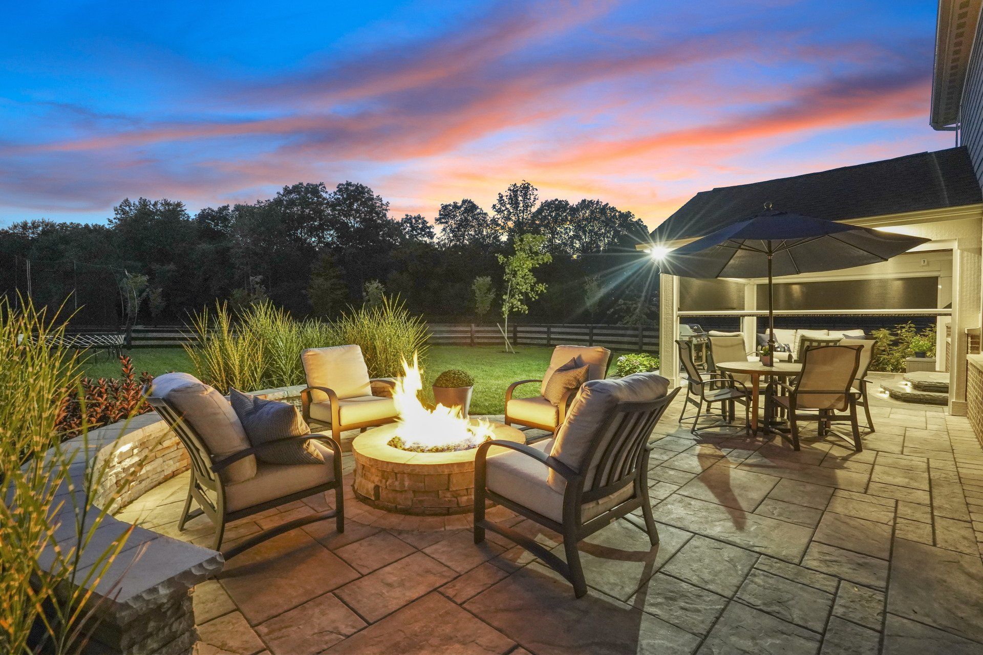 Backyard patio at dusk with fire pit, cushioned chairs, and a lit dining area under a covered porch