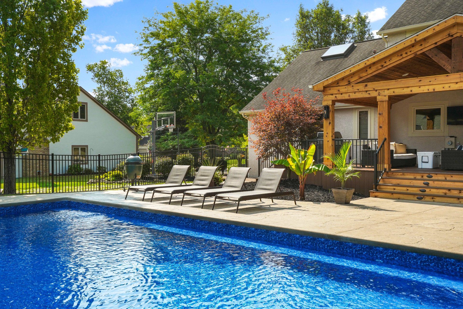 Backyard pool with lounge chairs beside a wooden patio and house in bright sunlight