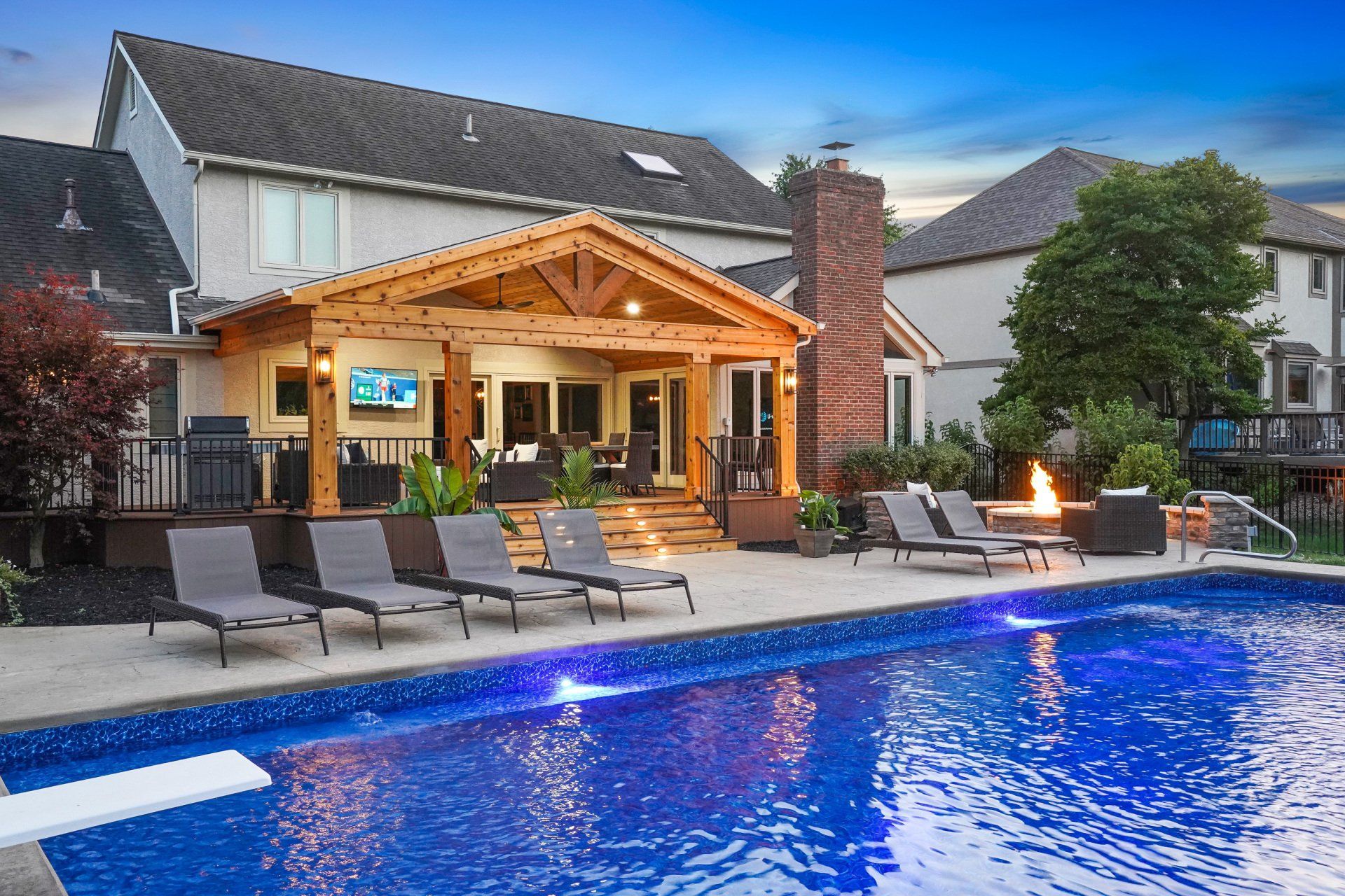 Backyard patio with lit pool, lounge chairs, fire pit, and a covered outdoor seating area at dusk