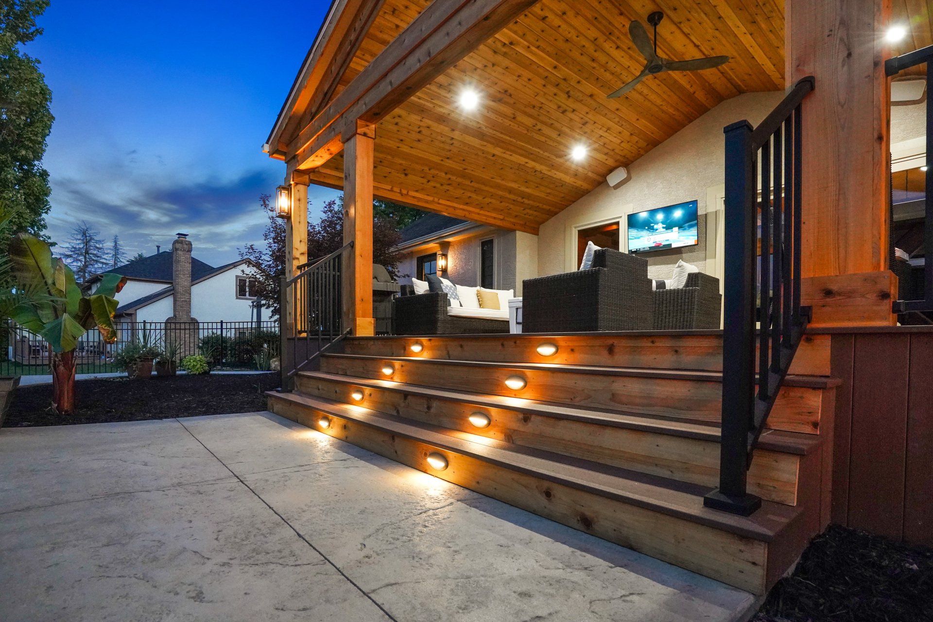 Warmly lit wooden porch with steps, black railings, and patio seating at dusk