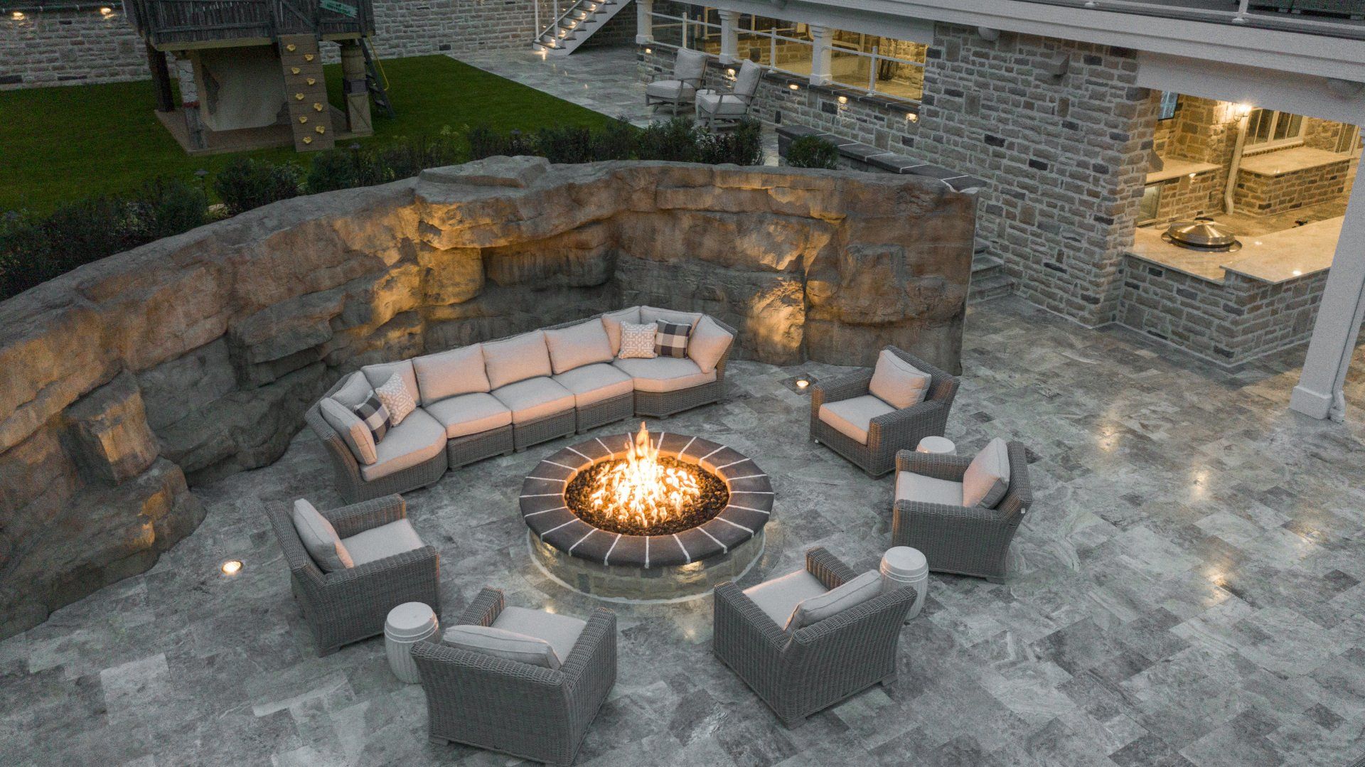 Stone patio fire pit with curved gray sofas and chairs arranged around a lit central flame at dusk