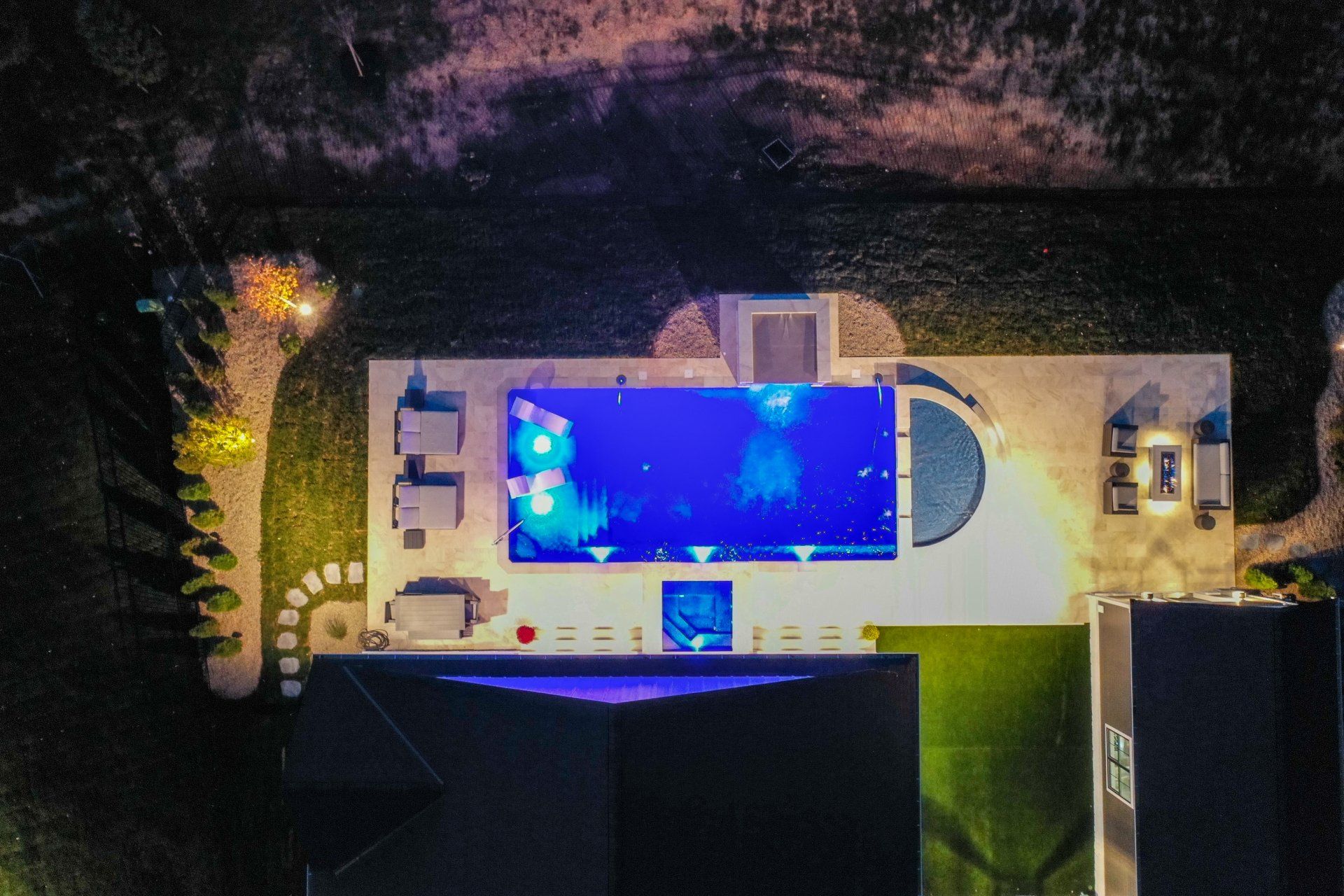 Aerial night view of a lit backyard with a rectangular swimming pool and surrounding patio.