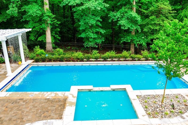 Backyard with a blue swimming pool, hot tub, and lush green trees surrounding the patio