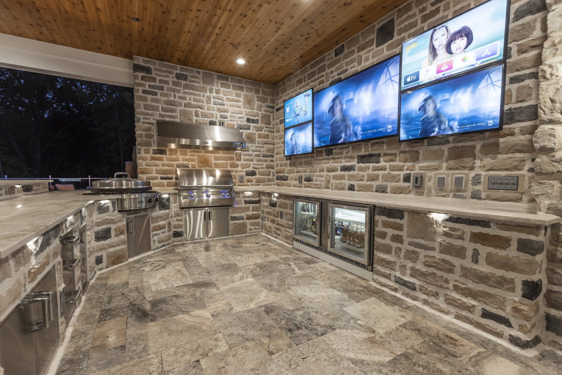 Stone outdoor kitchen with multiple wall-mounted TVs and stainless steel grill under a wooden ceiling