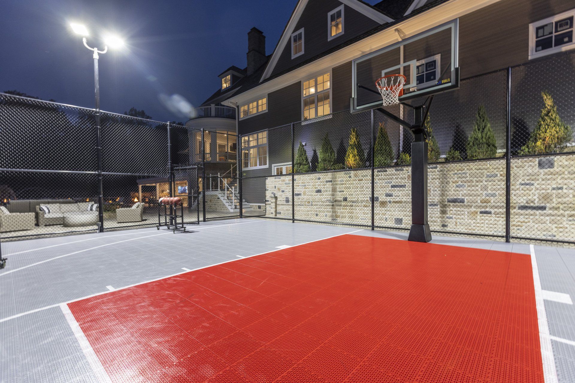 Lit outdoor basketball court with red-painted key, hoop, and stone wall beside houses at dusk