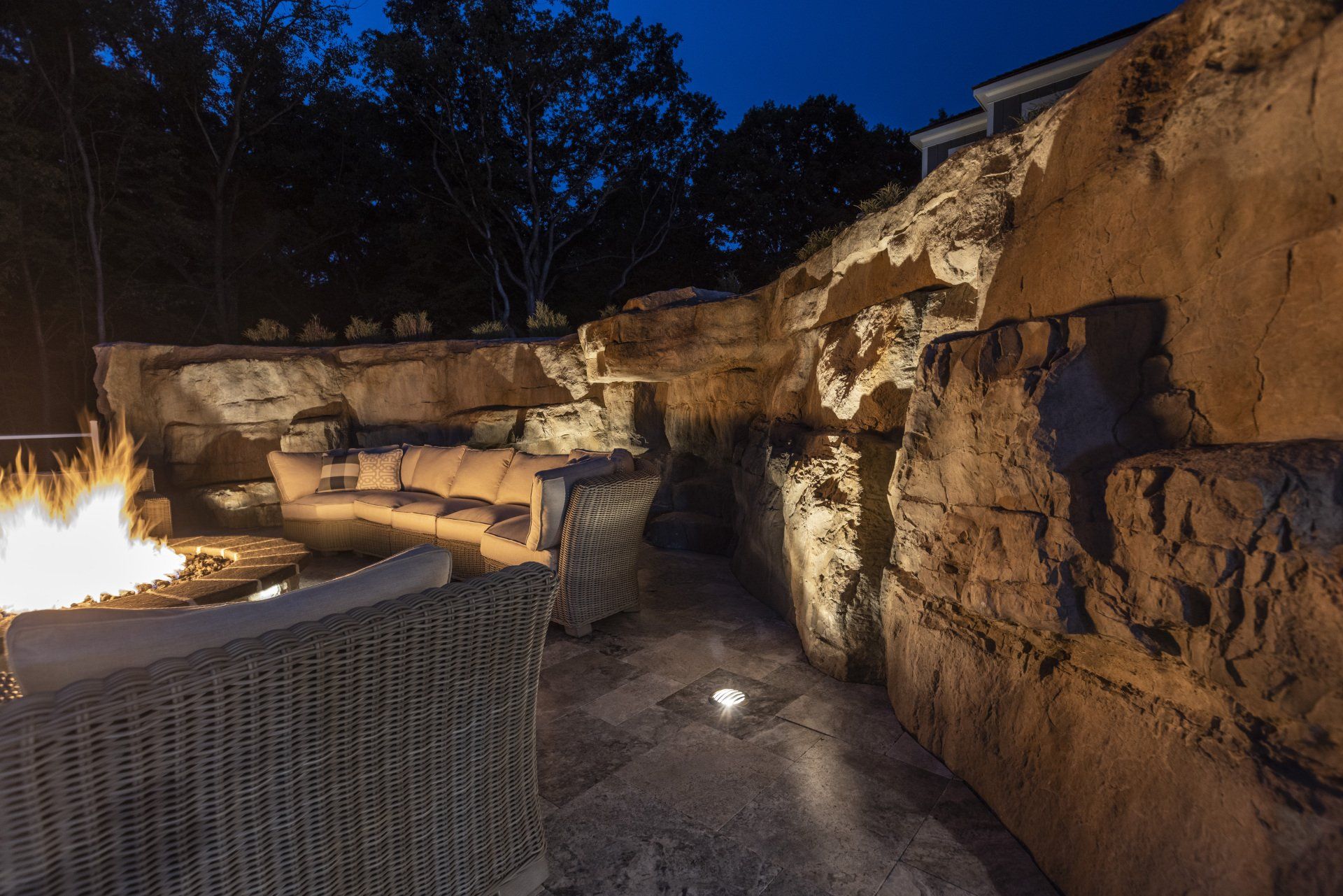 Stone patio with glowing fire pit, wicker chairs, and lit rock wall at night