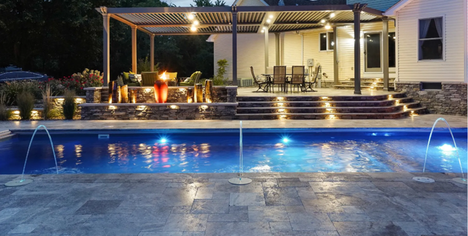 Blue-lit backyard pool at night with steps, patio seating, and a lit pergola beside a house