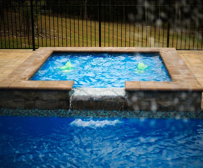 Small stone hot tub with blue water spilling into a larger blue pool outdoors