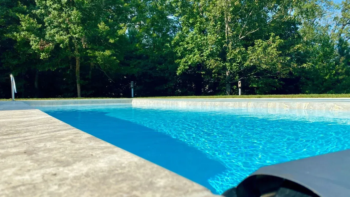 Bright blue swimming pool beside a concrete deck, with trees in the background.