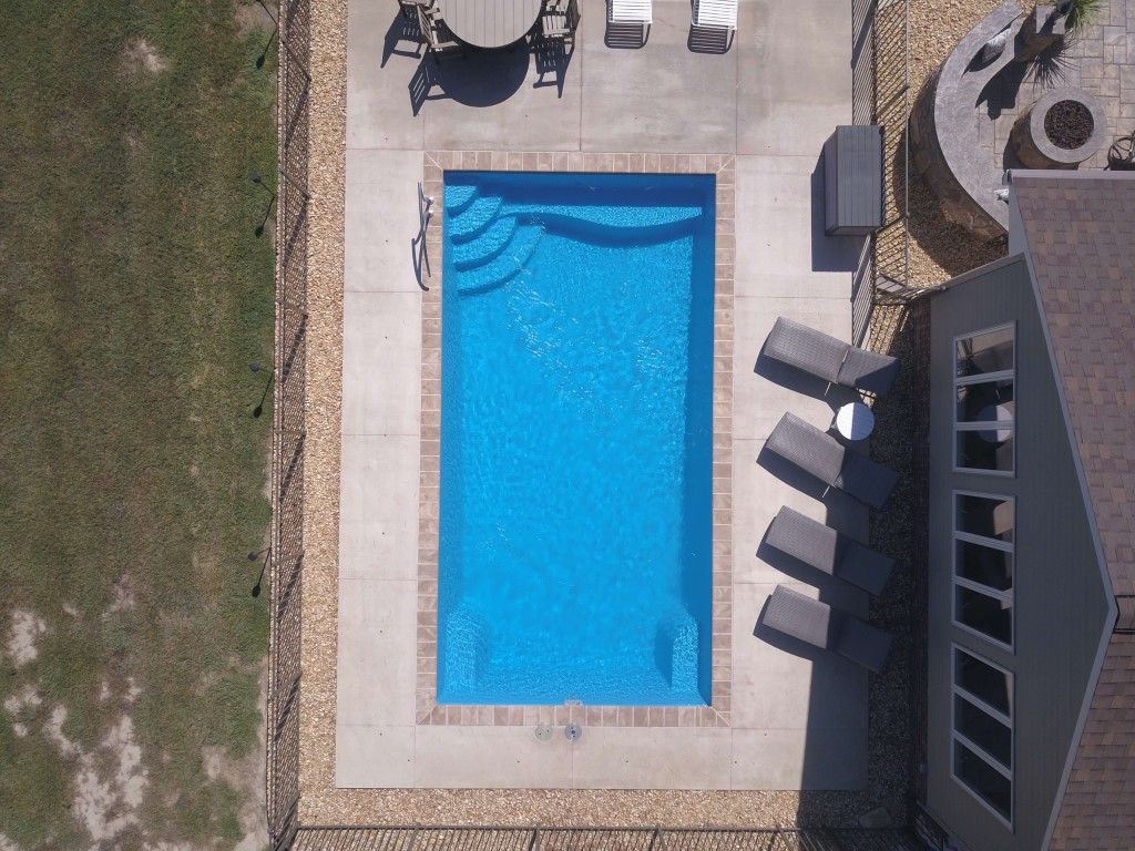 Aerial view of a rectangular blue swimming pool beside a patio with lounge chairs and a building.