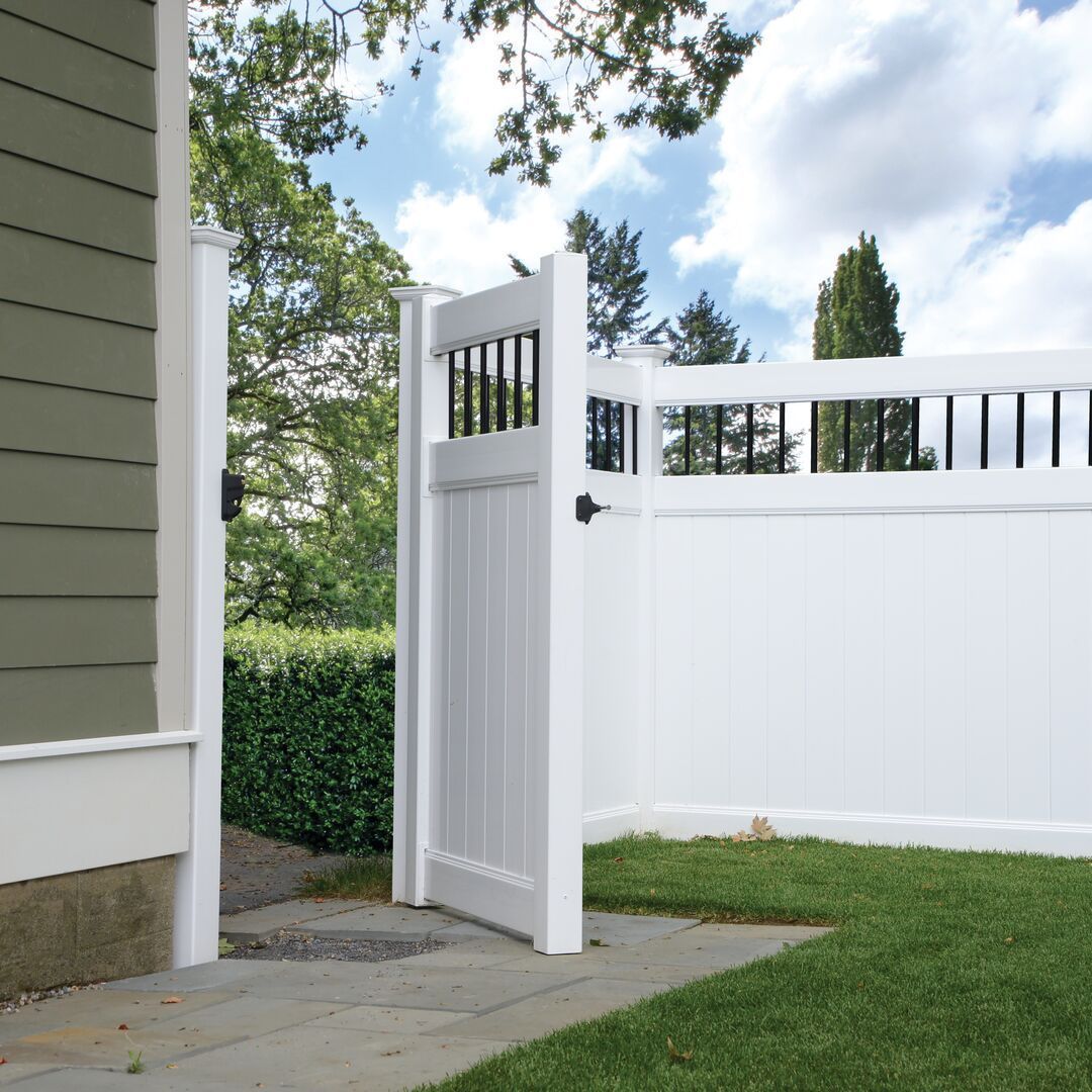 White backyard gate in a fenced lawn beside a house, opening onto a stone path under a partly cloudy sky