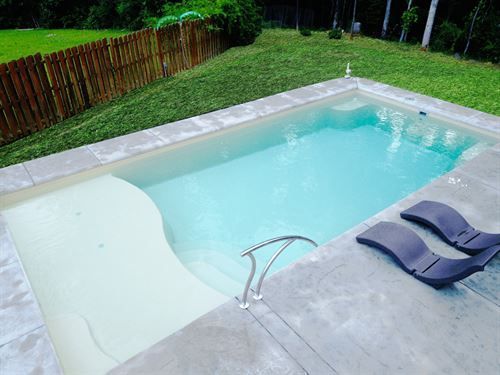 Backyard pool with light blue water, a shallow shelf, and two dark lounge chairs on the deck