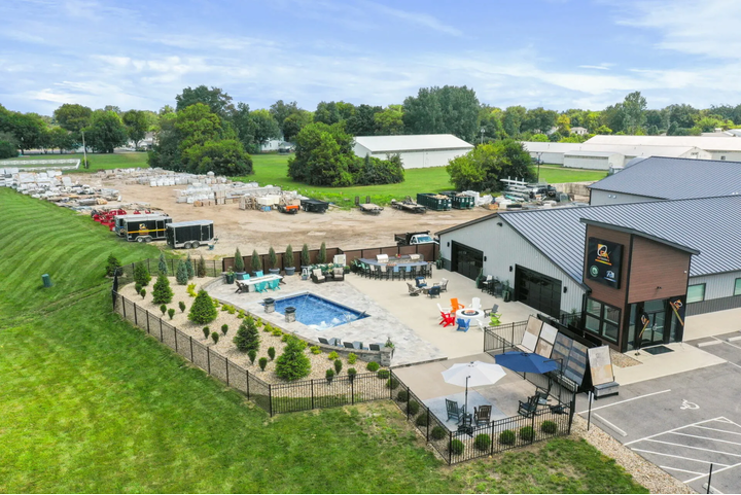 Aerial view of a landscaped patio with pool, outdoor seating, and a modern building beside green fields