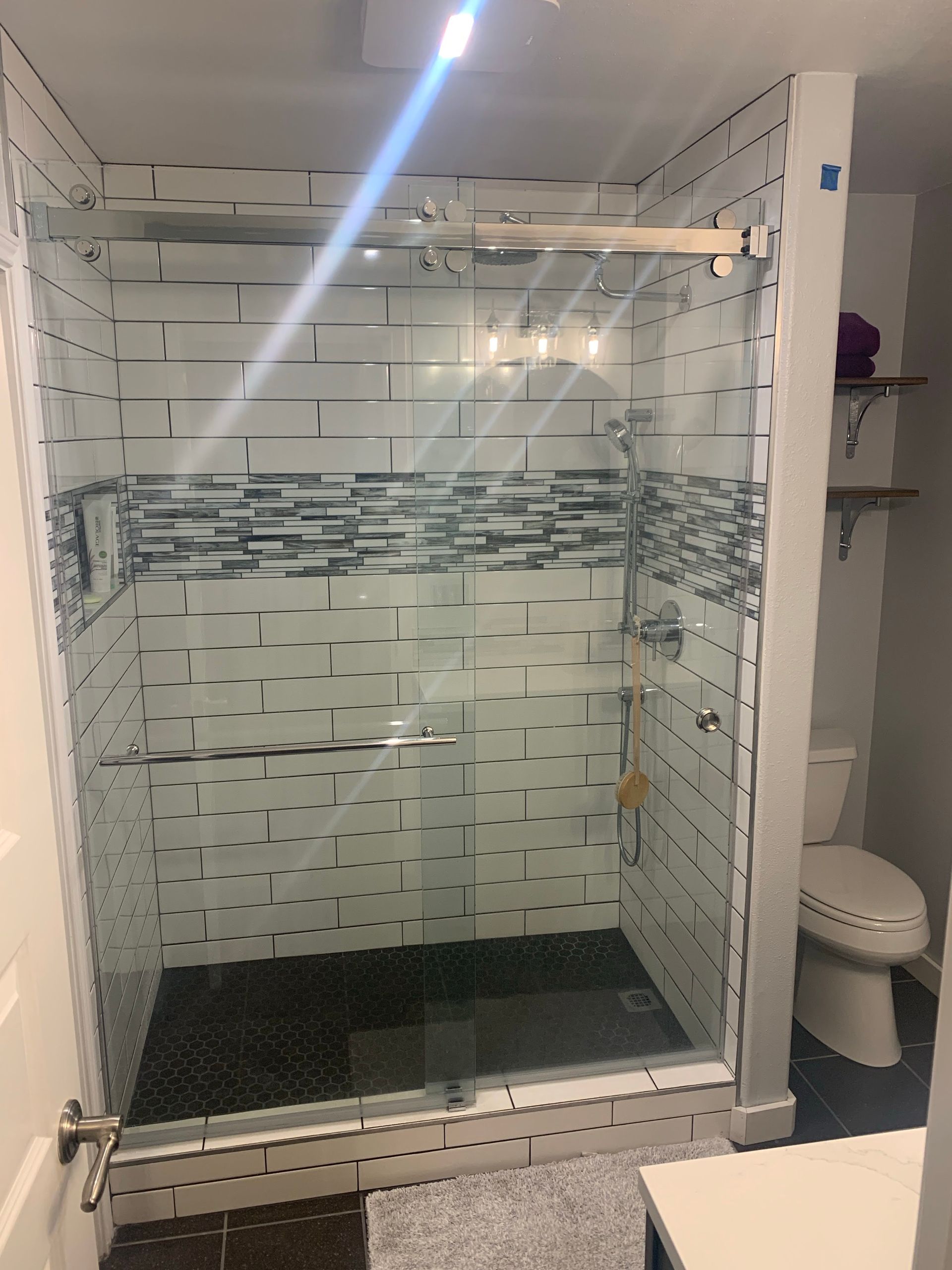Glass shower stall with mosaic tile accents and a dark floor mat, viewed from the bathroom doorway