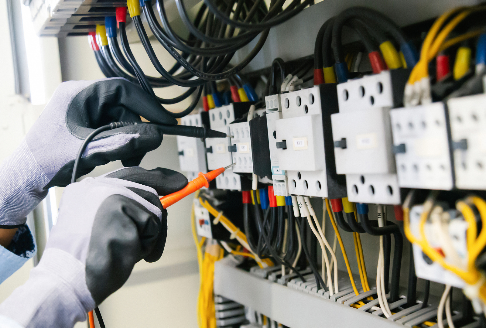 Hands in work gloves use multimeter probes to test connections on a circuit board inside an electrical panel.