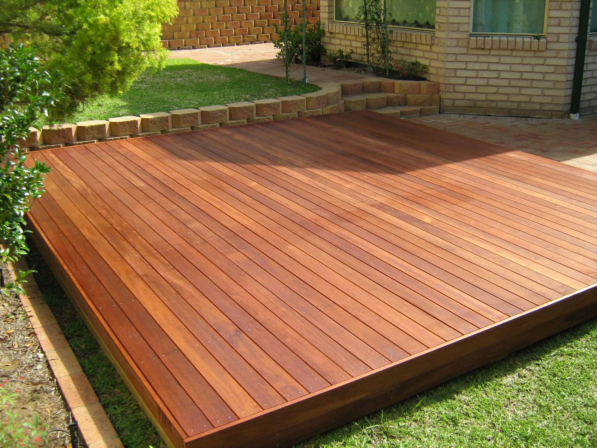 A rectangular, polished wooden deck set in a backyard next to a brick house and a small lawn.