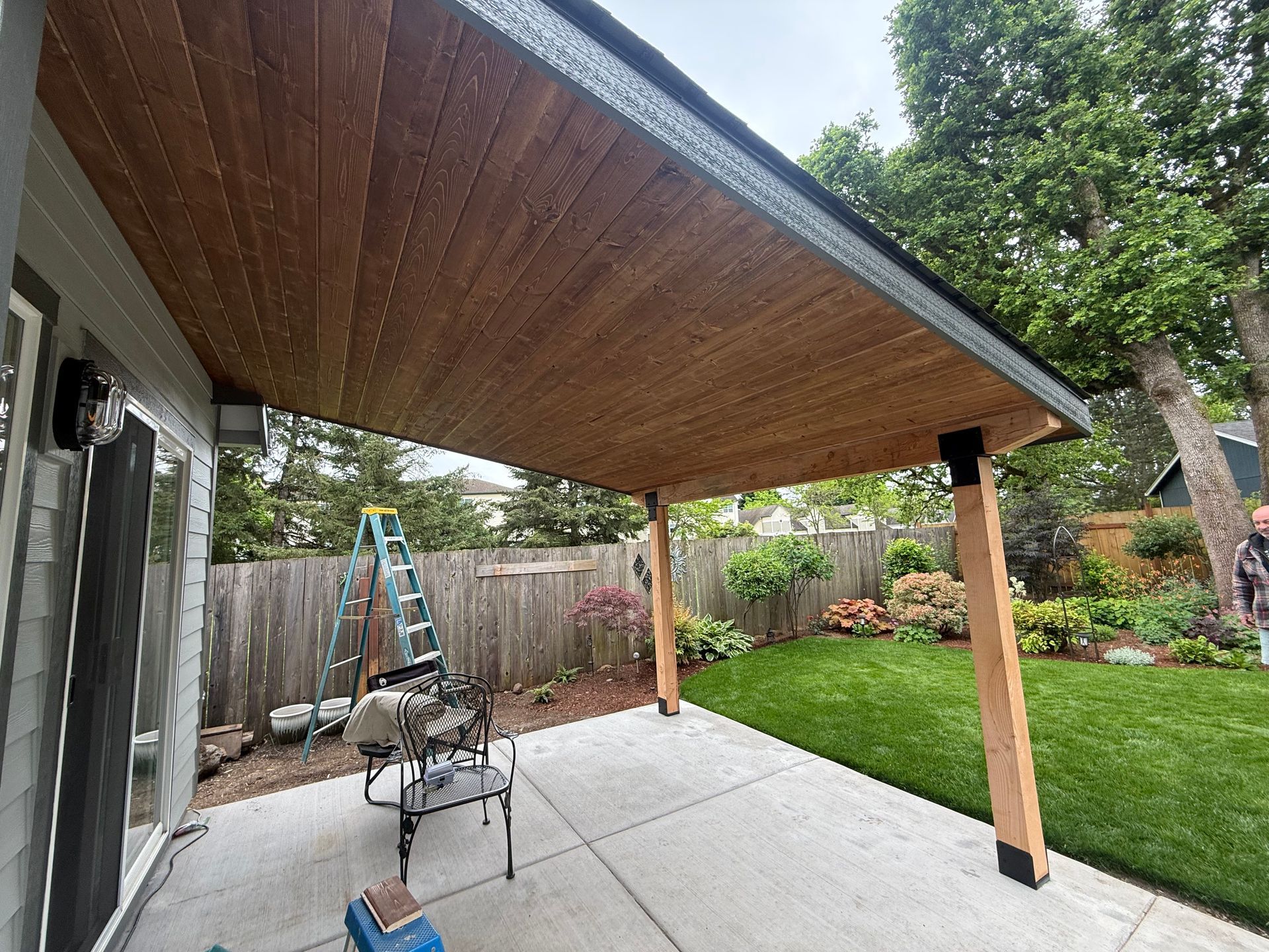 Covered patio with wooden ceiling, concrete floor, ladder, and outdoor seating in a backyard garden