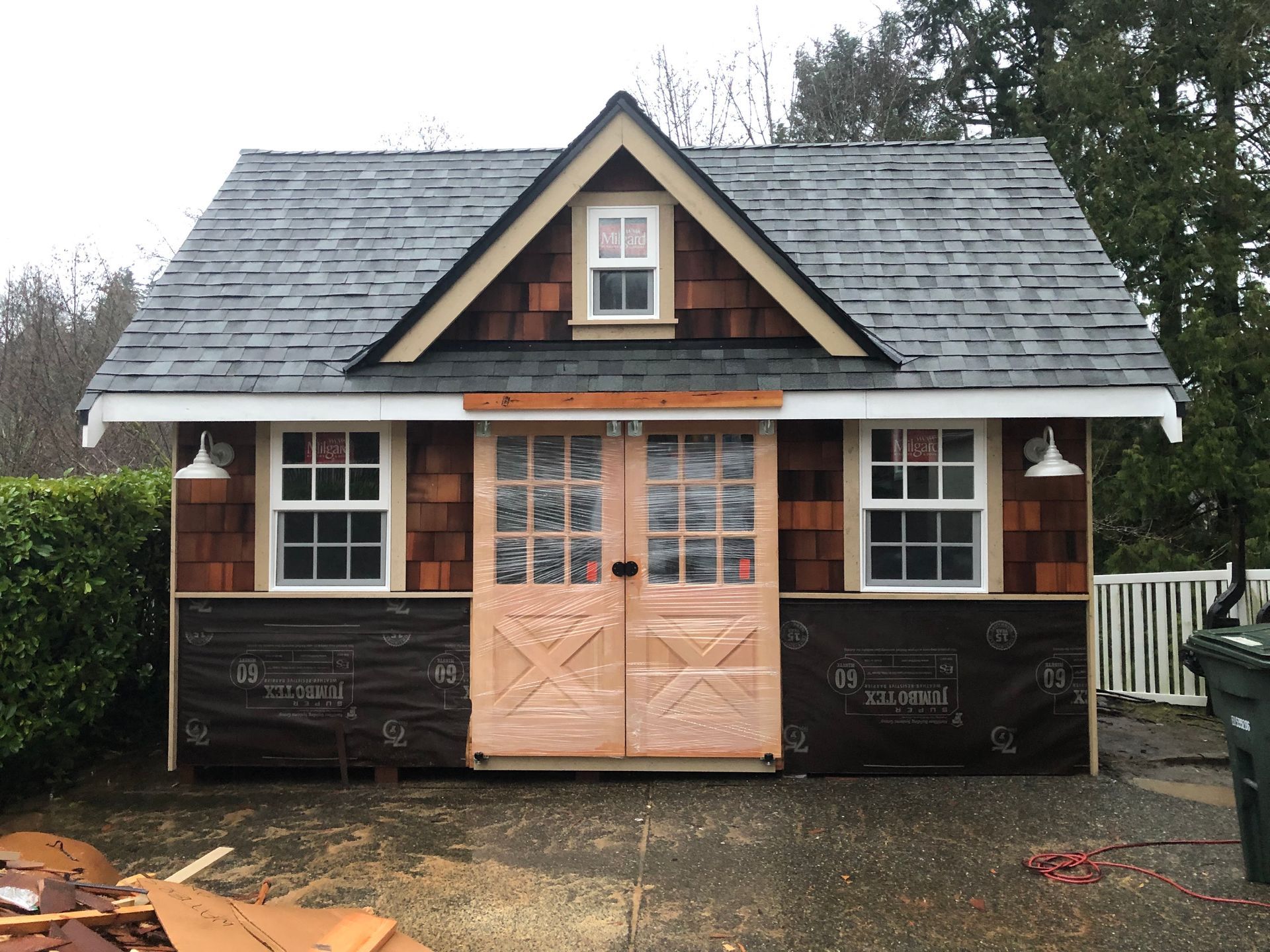 Small cottage-style shed with a tan double door, dormer window, and dark shingled roof in a yard.