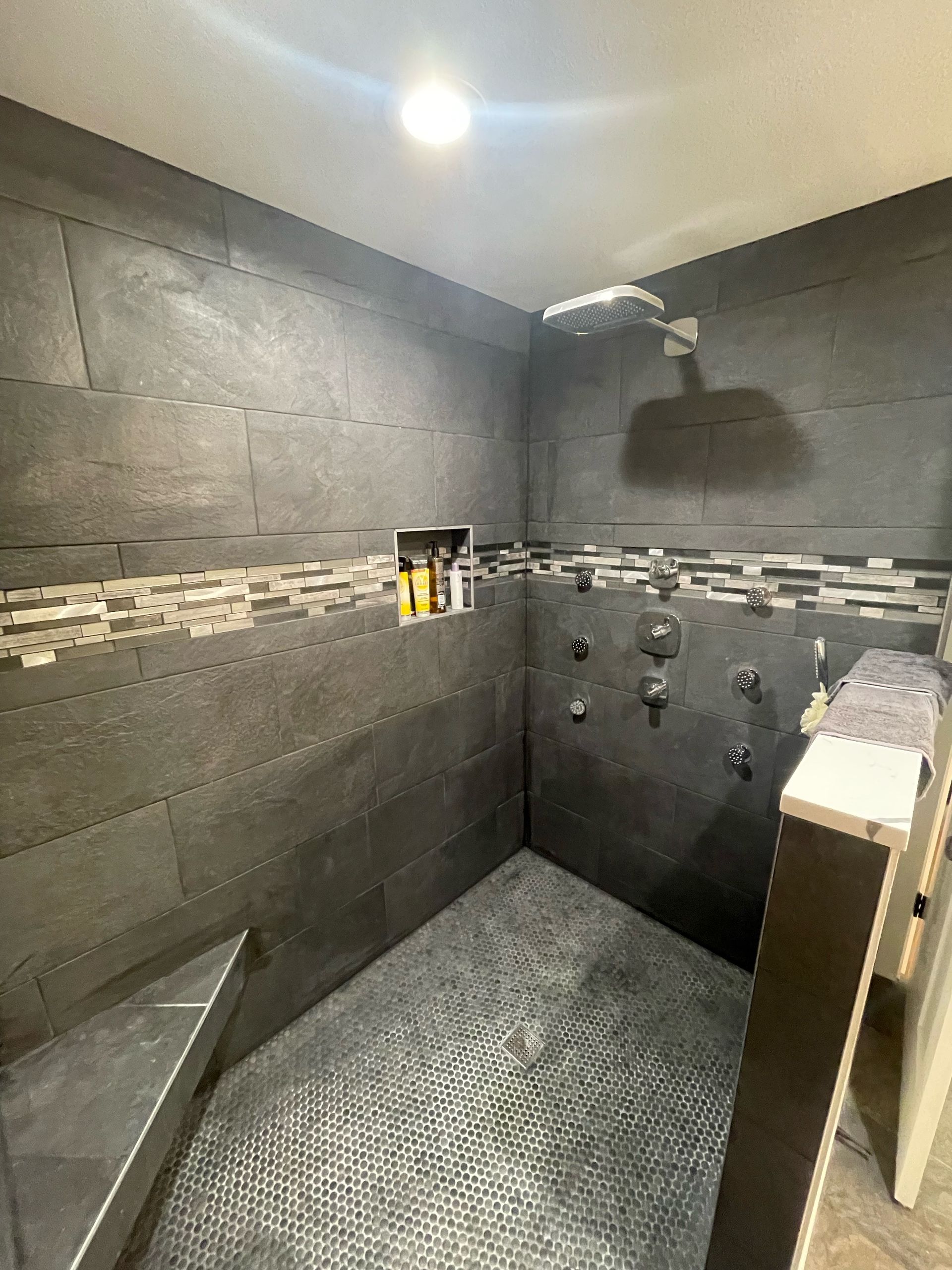 Modern shower with gray stone-tile walls, mosaic accent band, and multiple wall-mounted fixtures.
