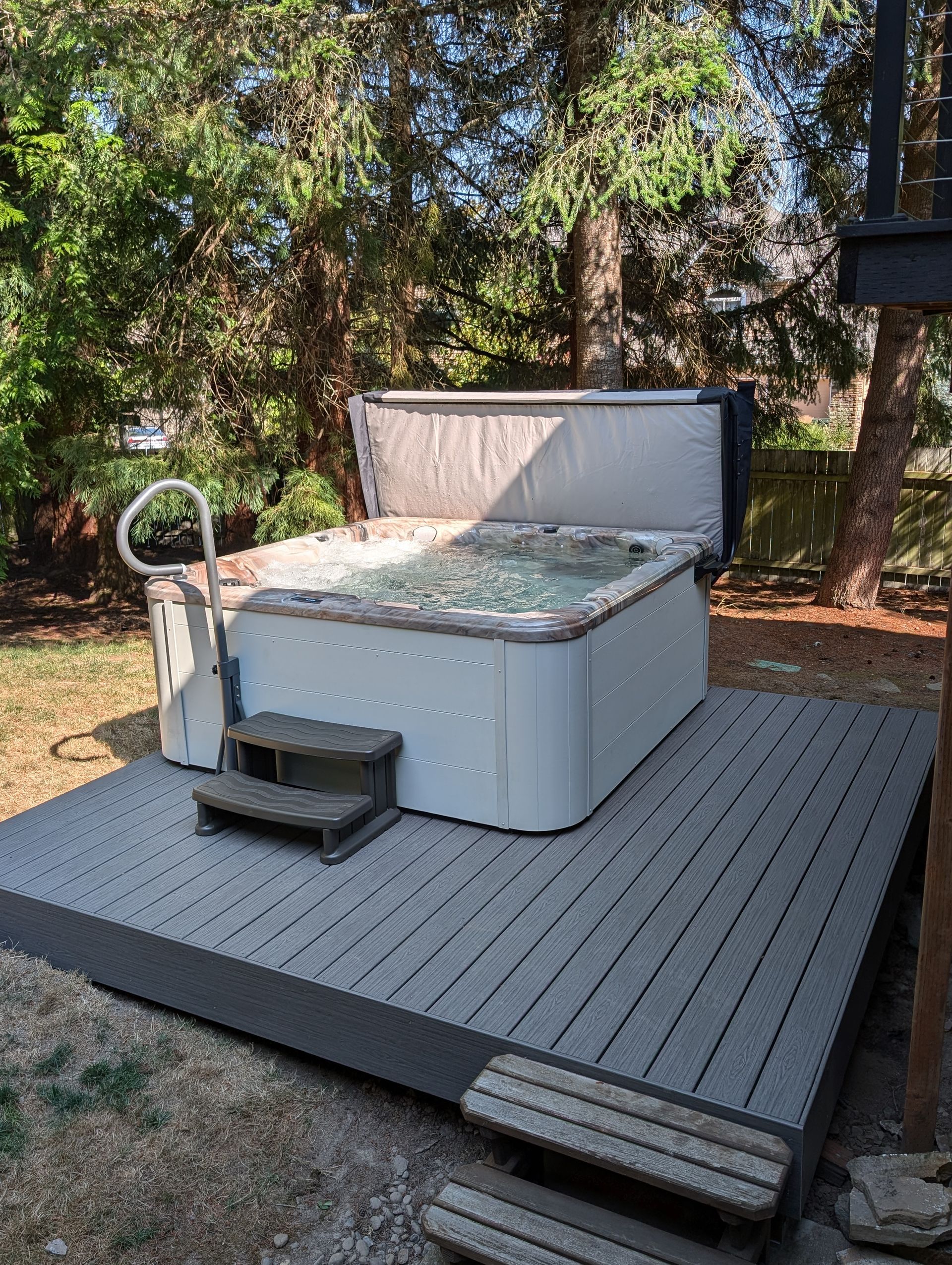 White hot tub on a gray deck in a wooded backyard, with a step and handrail.