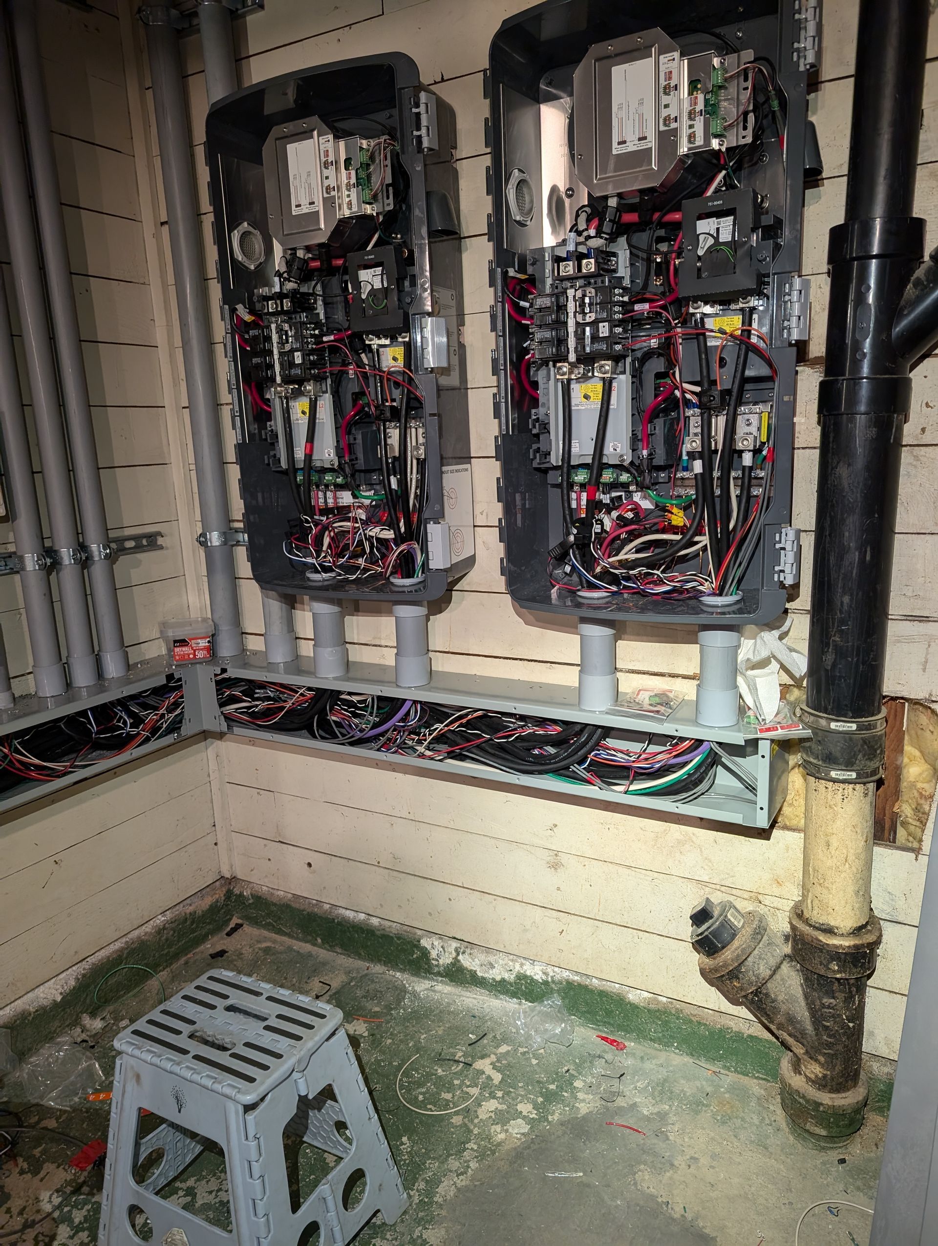 Open electrical panels and exposed wiring in a utility room, with pipes and a small step stool.
