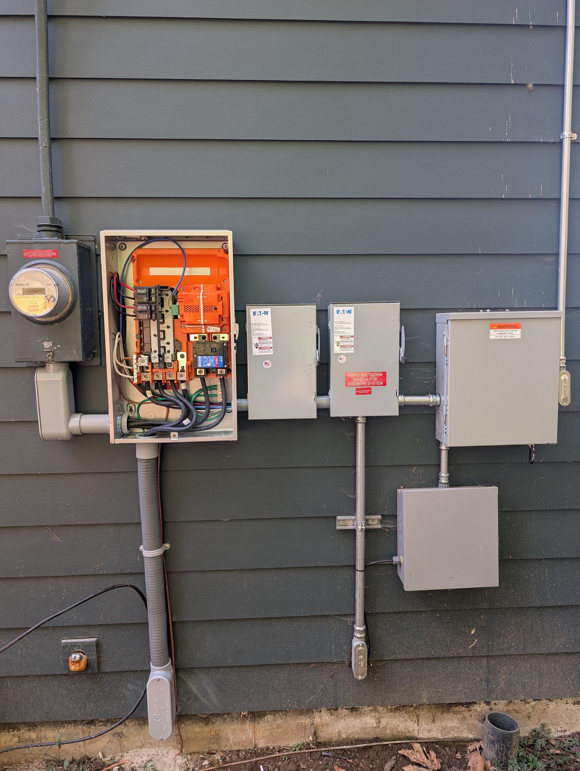 Electrical meter and utility boxes mounted on a gray exterior wall with exposed wiring