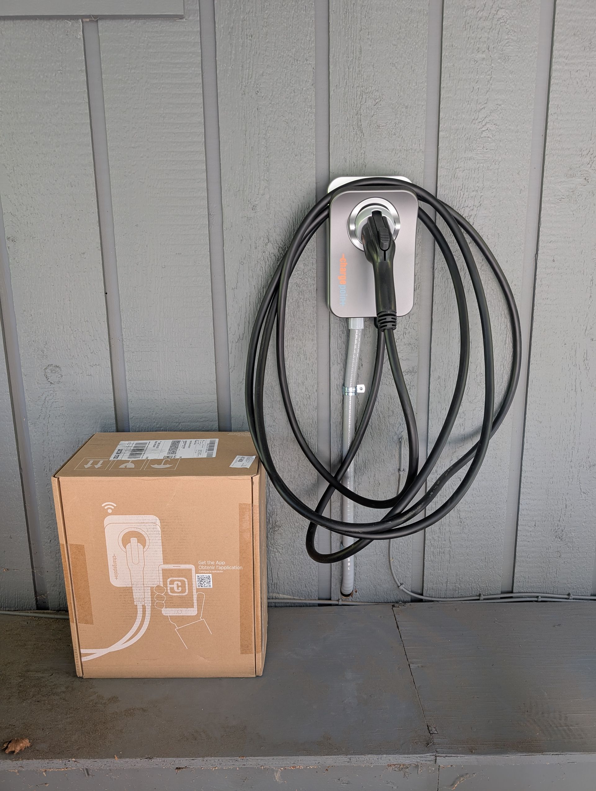 Cardboard box beside a wall-mounted charging station with a coiled black cable on gray siding.