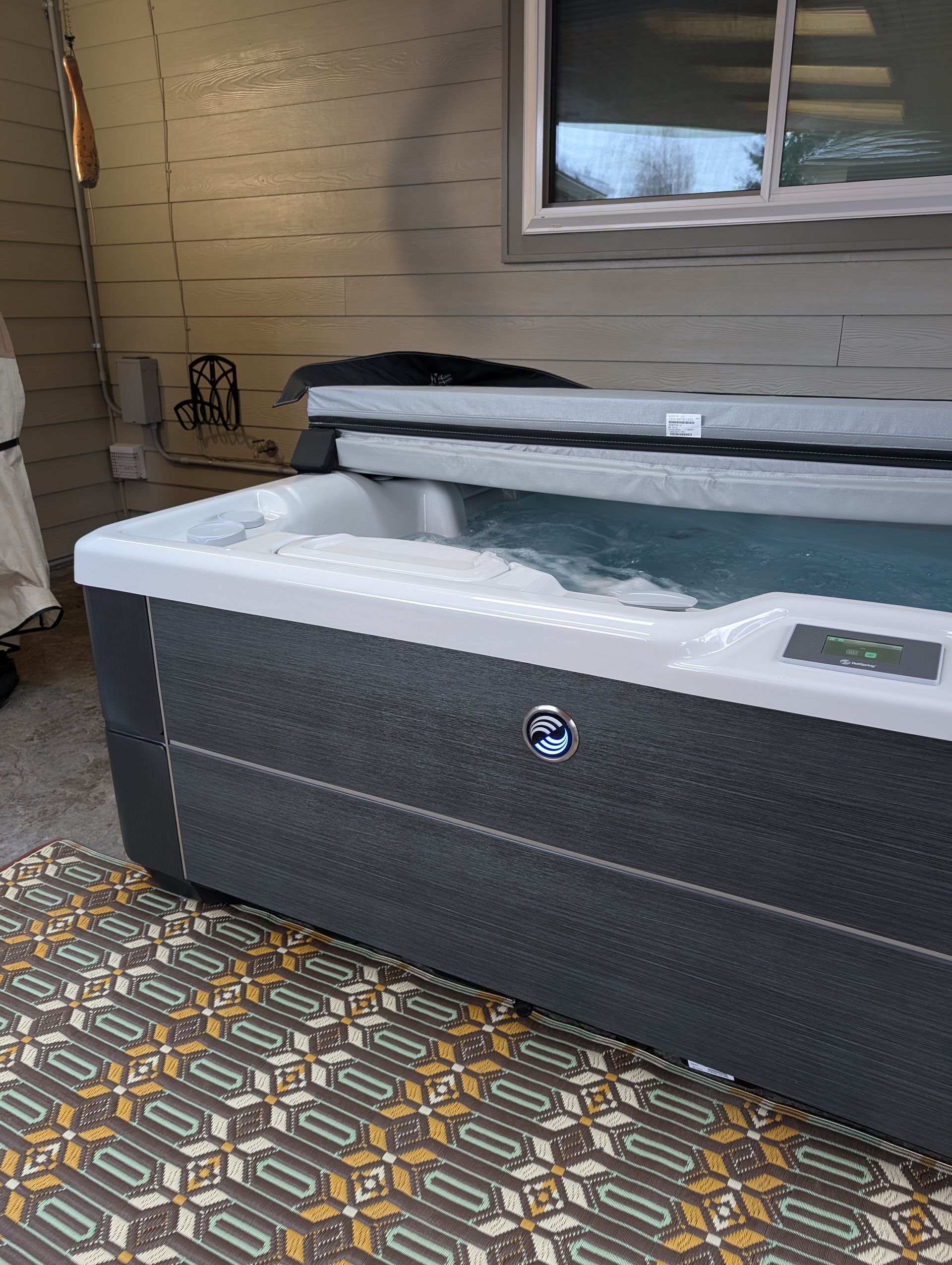 Gray hot tub with open lid on a patterned patio beside a beige house wall and window.