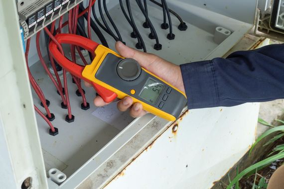 A person uses a yellow and black clamp meter to measure the electrical current on a red wire inside a control panel.