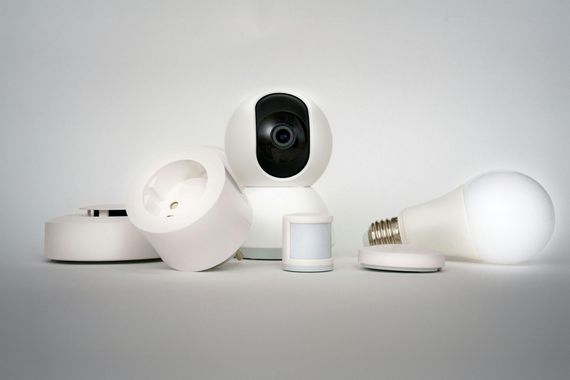 A collection of white smart home devices, including a camera, light bulb, power plug, and sensors, on a light background.