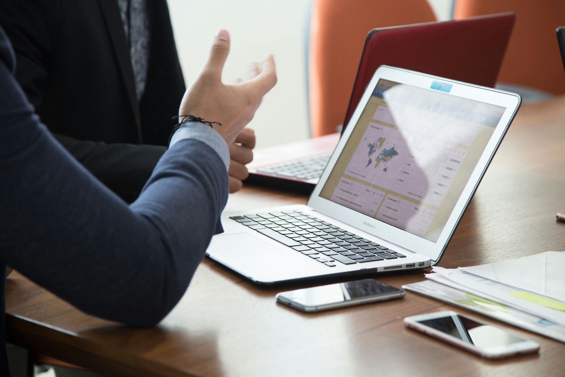 Two people at a table, discussing data on a laptop. There are cell phones and a second laptop present.