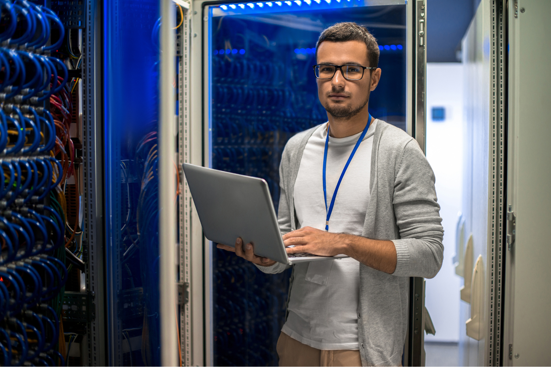 Man with laptop in a server room, looking at the camera. He wears glasses, a lanyard, and a light gray cardigan.