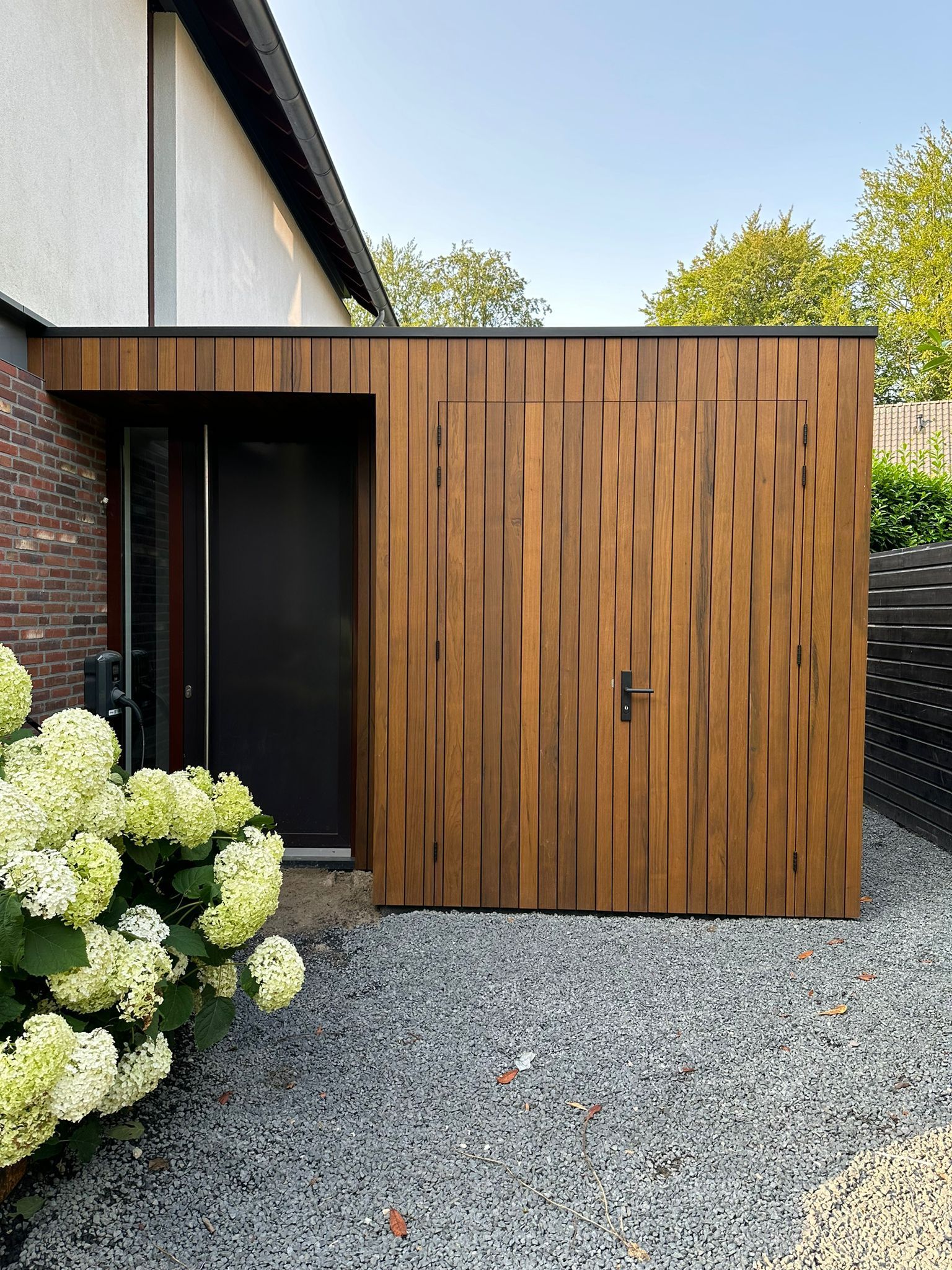 Moderne woning met houten poort, grindoprit, bakstenen muur en hortensiastruiken.
