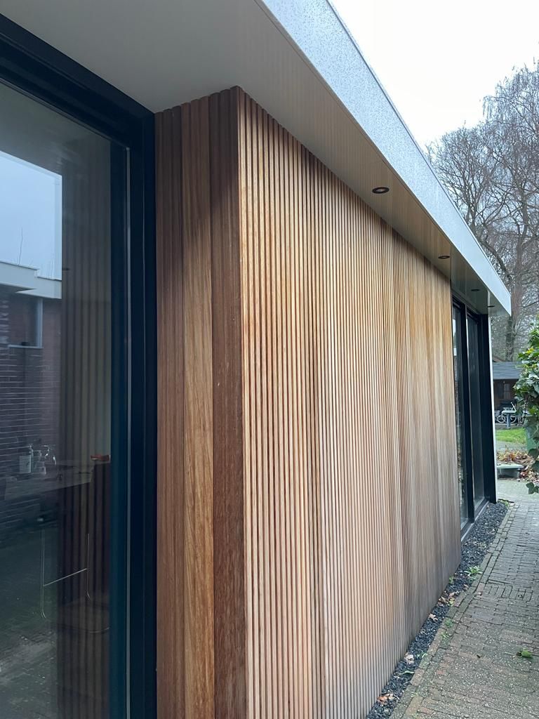 Een met hout beklede buitenmuur naast een glazen deur, met een grindpad en bomen op de achtergrond.