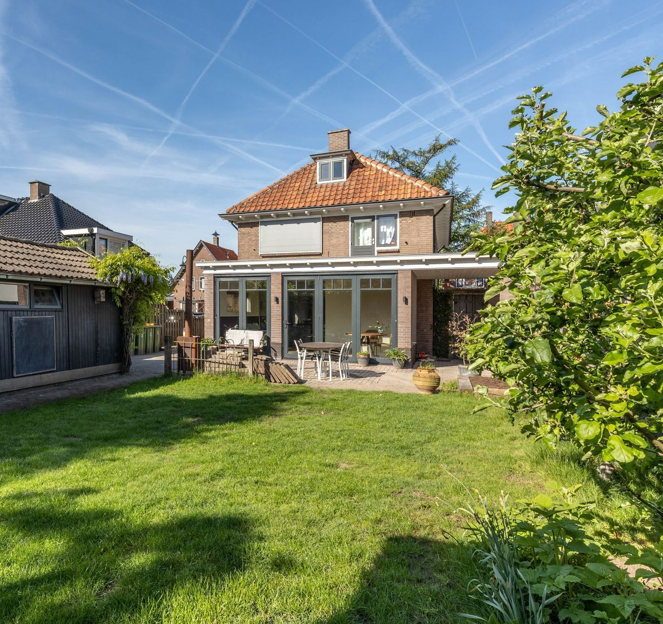 Achtertuin met een huis van twee verdiepingen, terrasmeubilair en een groot groen gazon onder een helderblauwe hemel.