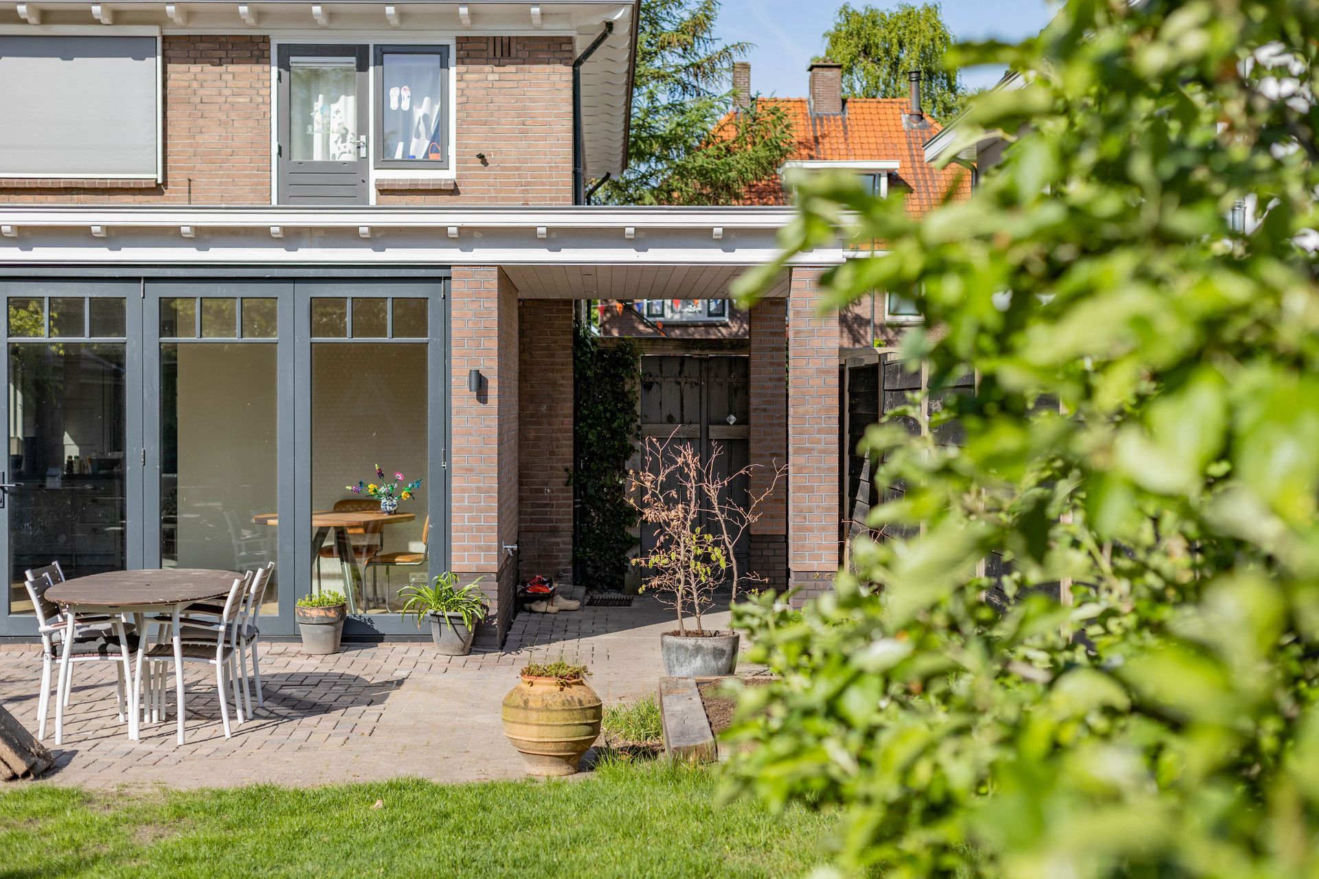 Terras in de achtertuin met een stenen huisje, glazen deuren, tafel, stoelen en potplanten onder een overdekte luifel.
