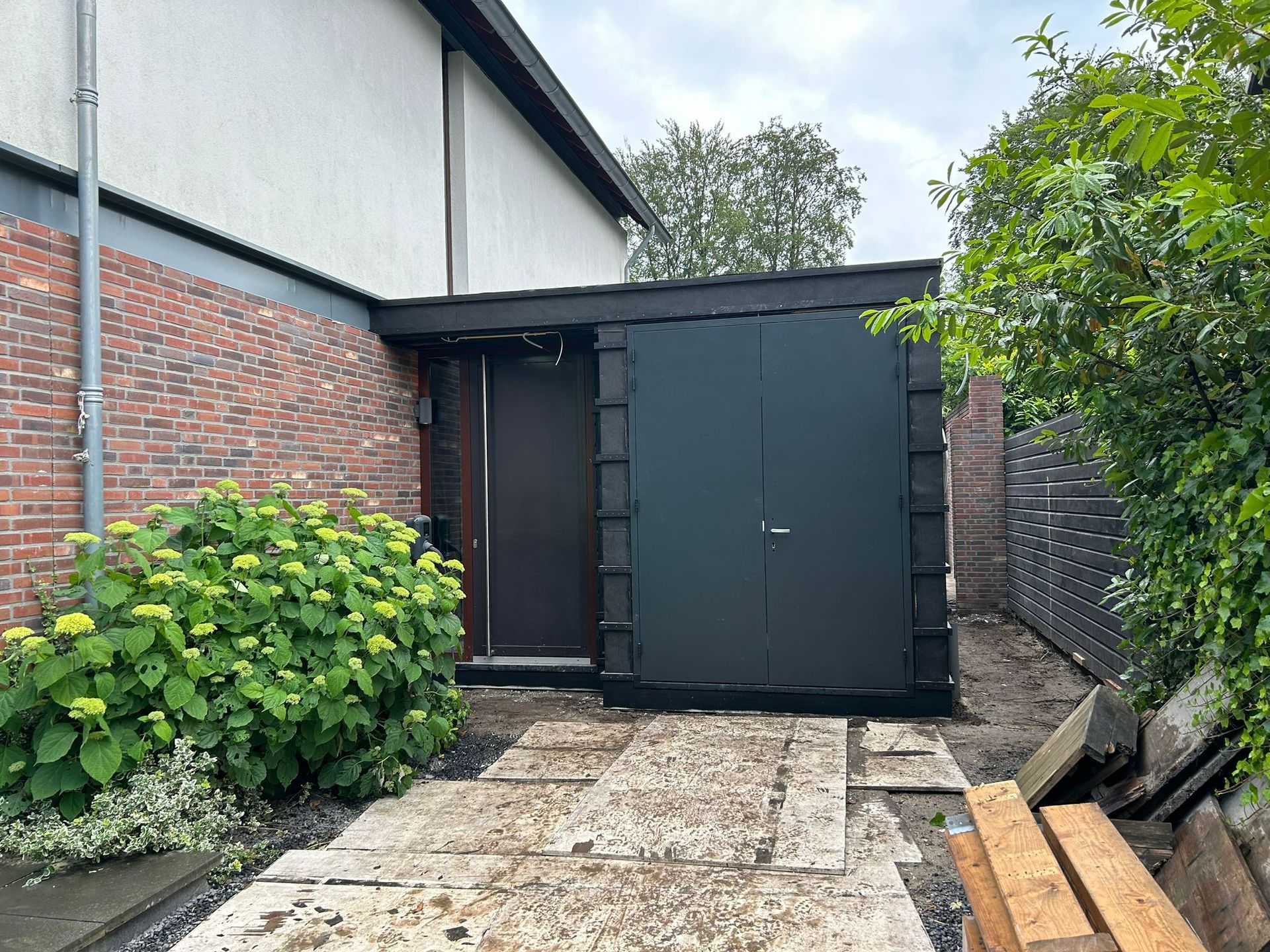 Terras in de achtertuin met een donkere schuur, een bakstenen muur en stenen bestrating, omgeven door groen.