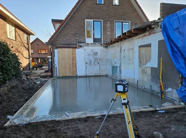 Een verse betonnen plaat is gestort tijdens een tuinrenovatie, met een driepoot en onafgewerkte muren naast een huis.