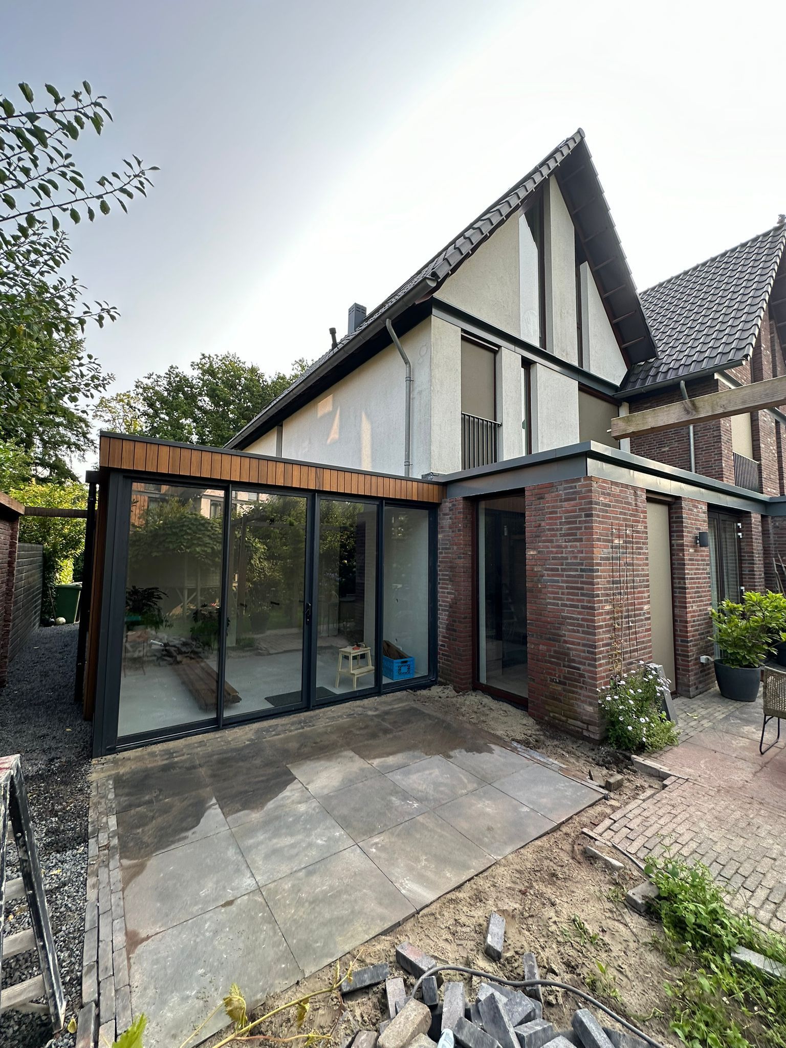 Glazen serre-aanbouw naast een bakstenen huis met een schuin dak en terrasbestrating.