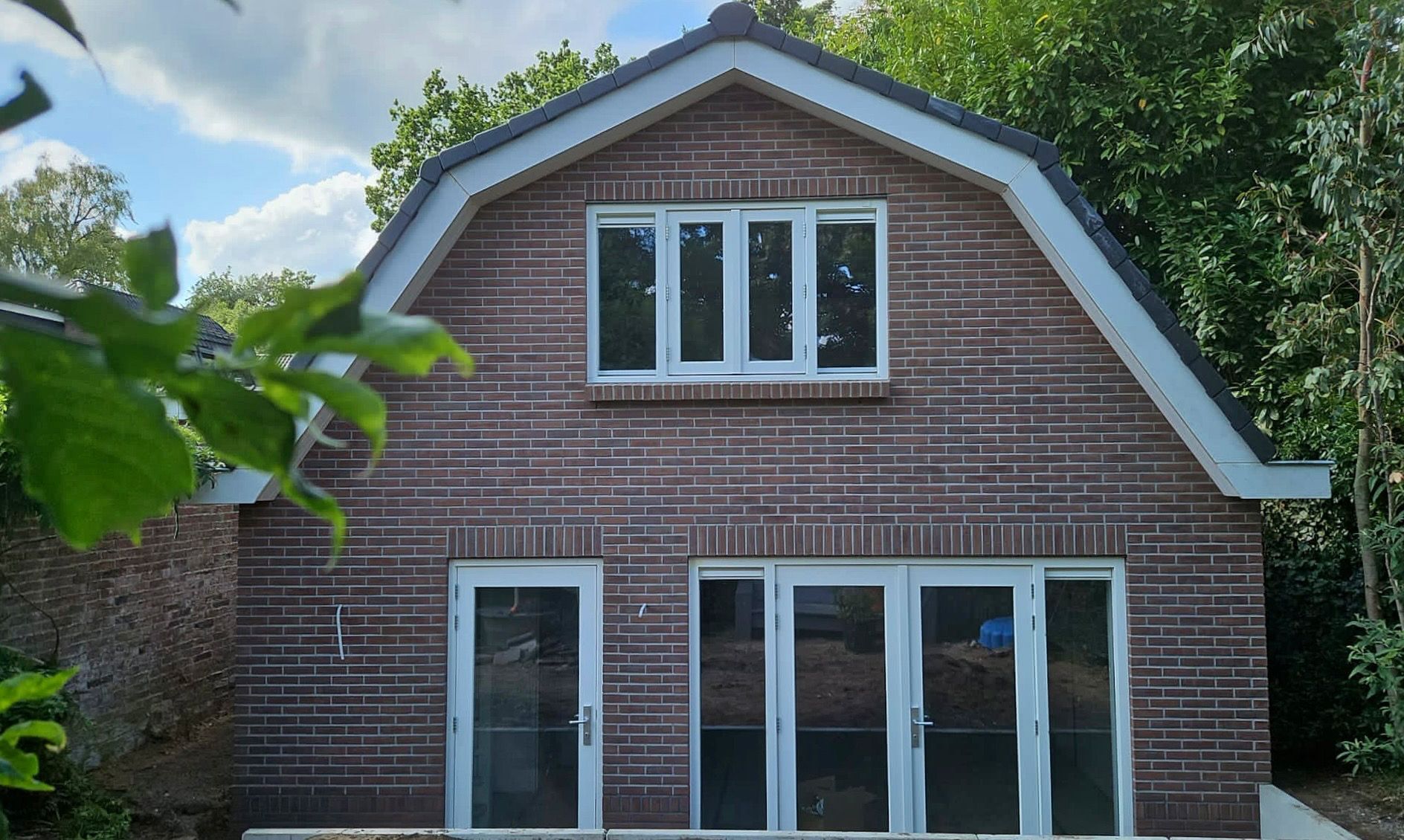 De buitenkant van een bakstenen huis met een zadeldak, witte kozijnen en grote glazen deuren.