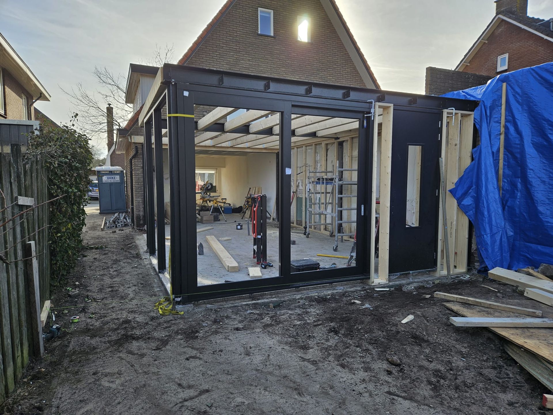 Gedeeltelijk gebouwde aanbouw met grote glazen deuren, puin en een blauw zeil in een modderige achtertuin.