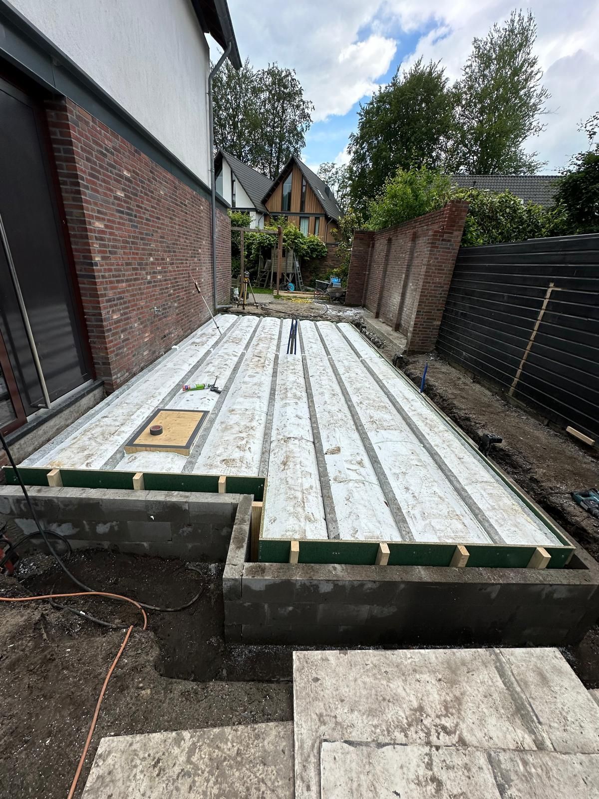 Een gedeeltelijk aangelegd houten terras naast een bakstenen huis, met planken over balken en een tuinpad in de buurt.