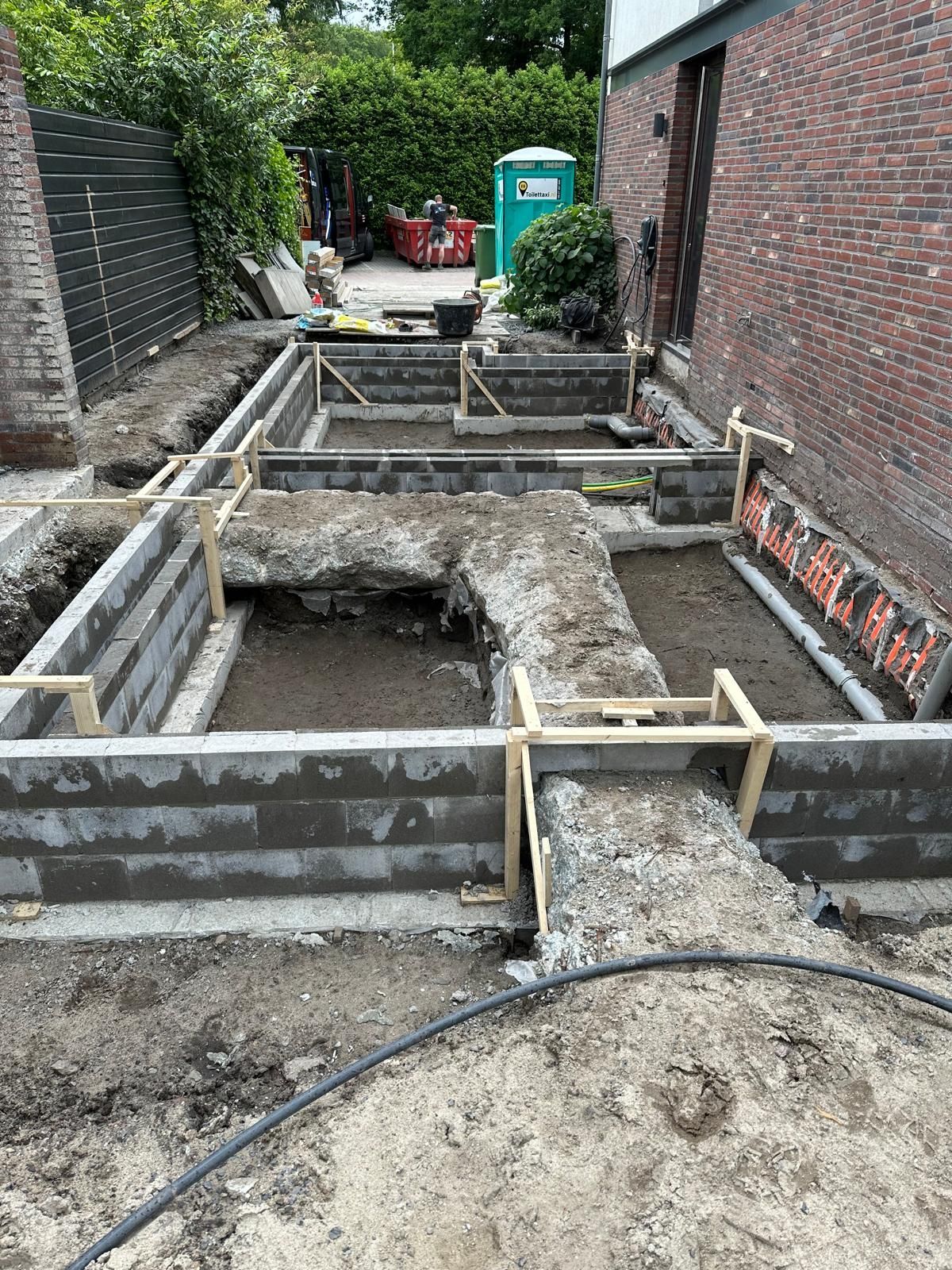 Bouwsleuffundering met houten bekisting en betonnen funderingsvoet naast een bakstenen muur