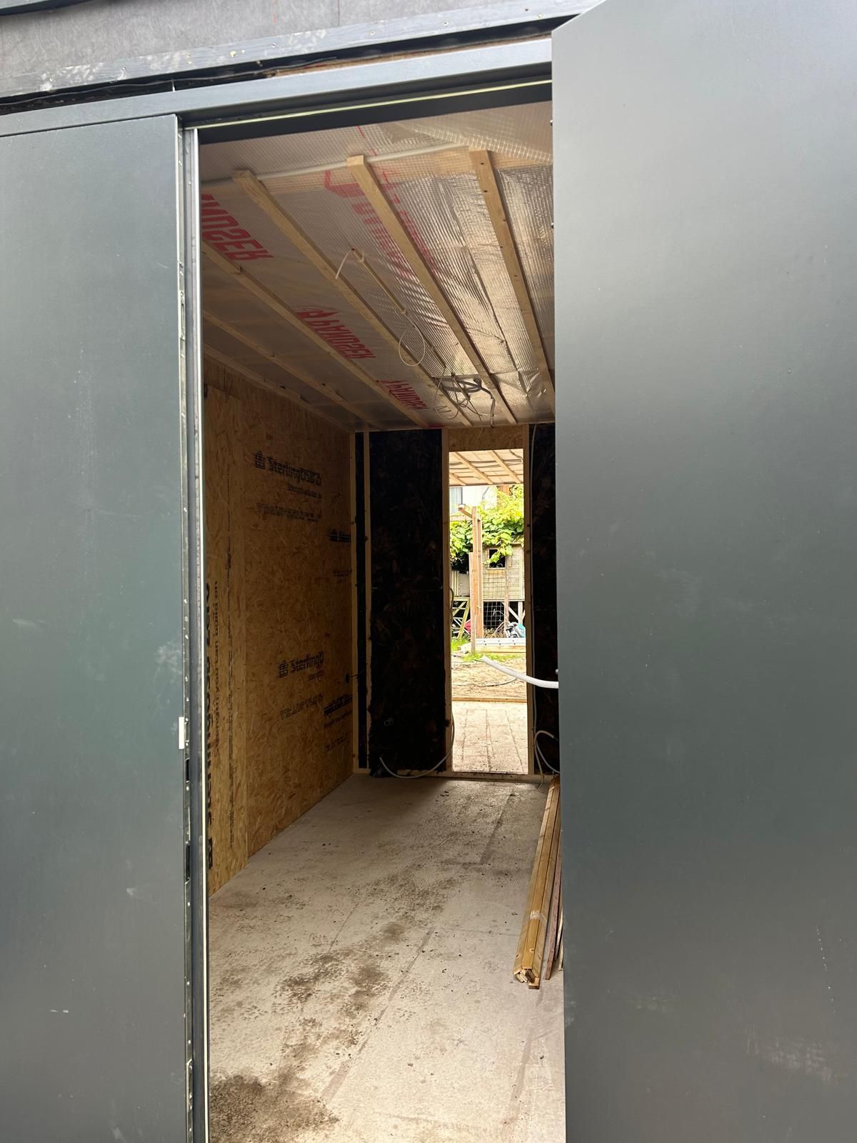 Open deuropening naar een onafgewerkte binnengang met zichtbare isolatie en multiplexwanden.
