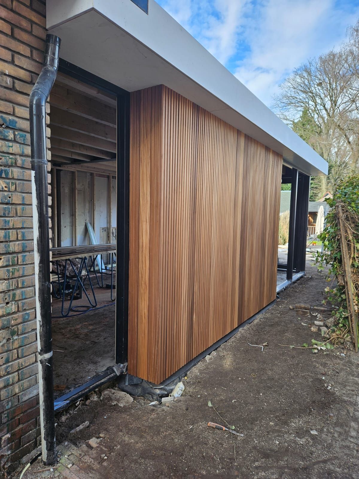 Houten schuur of muur naast een huis, onder een schuin dak, langs een smal buitenpad.