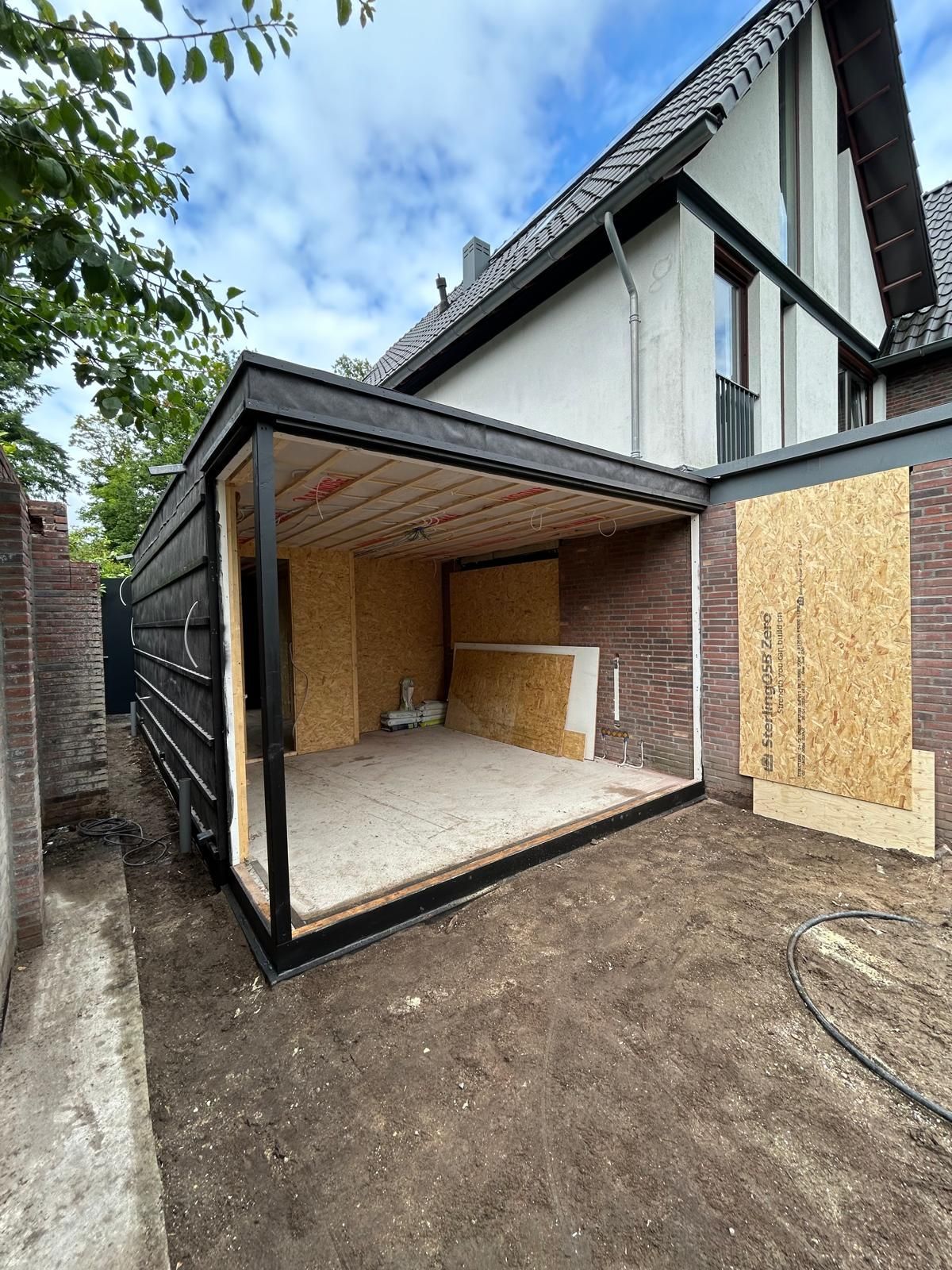 Open garage/carport in aanbouw naast een huis, met onafgewerkte houten wanden en een ondergrond van aarde.