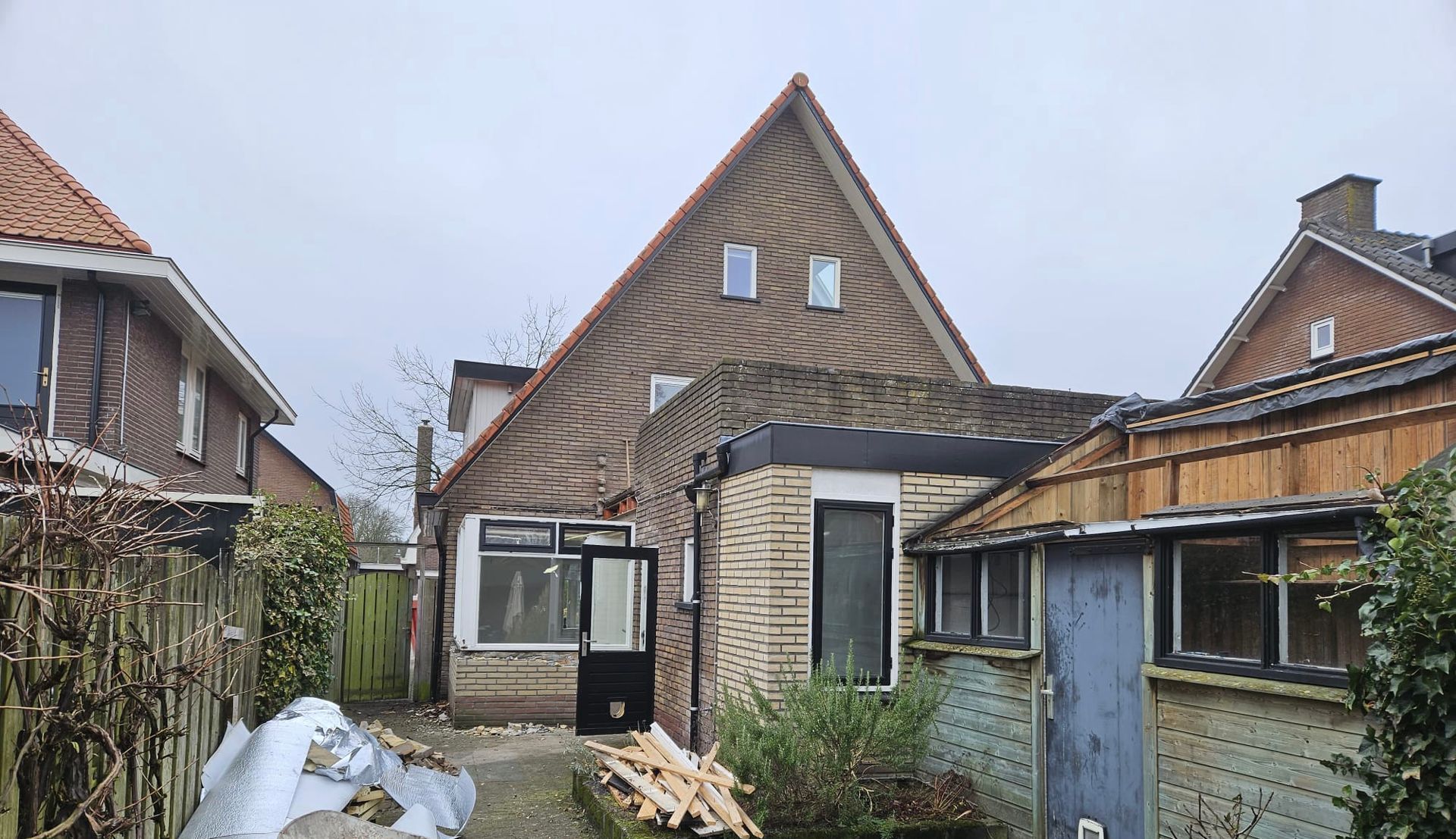 Achtertuin met een bakstenen huis, schuur en smal pad, met tuingereedschap en sneeuw op de grond.