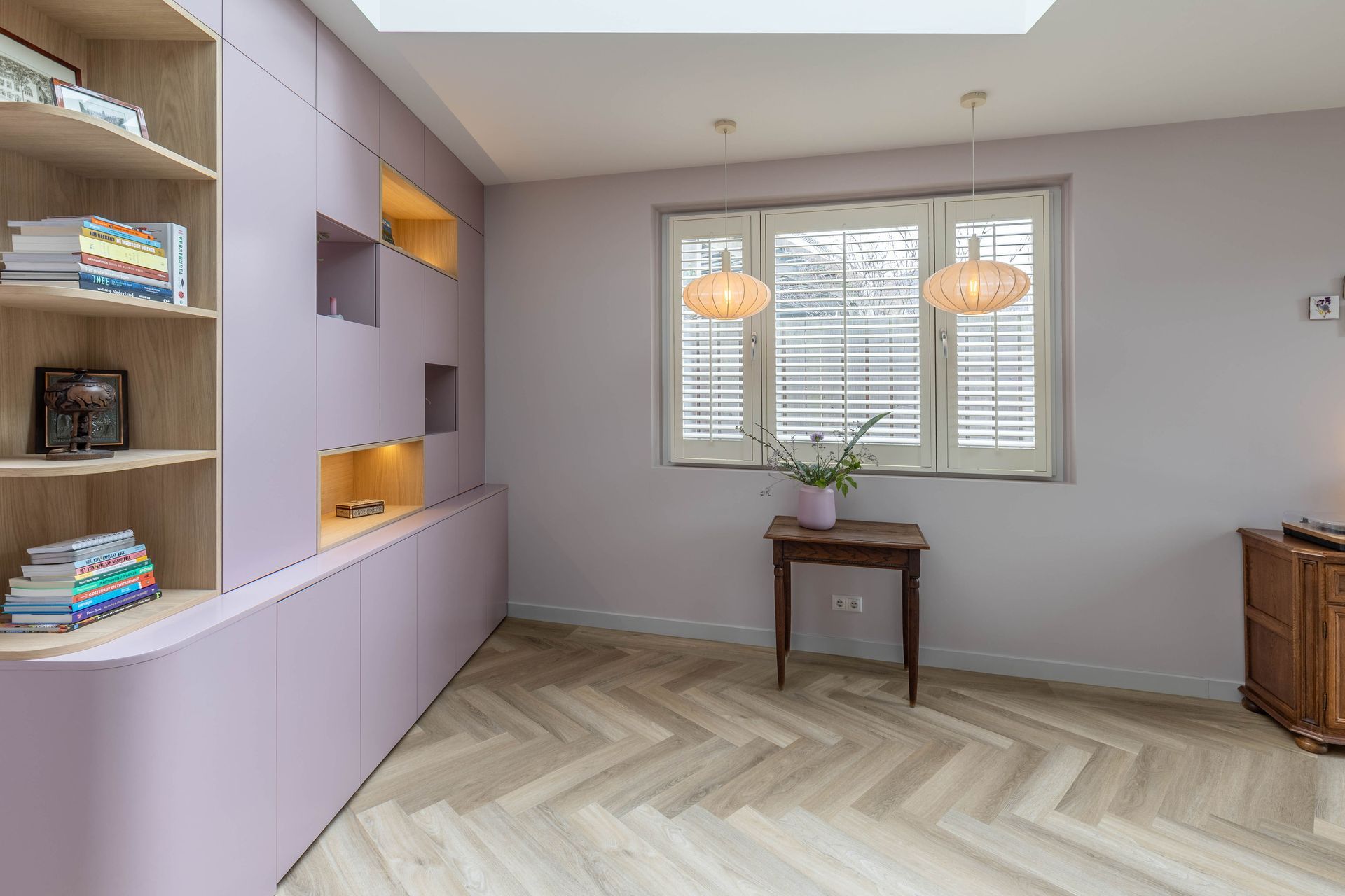 Licht lavendelkleurige kamer met ingebouwde planken, raamluiken en een klein tafeltje op een houten vloer in visgraatpatroon.