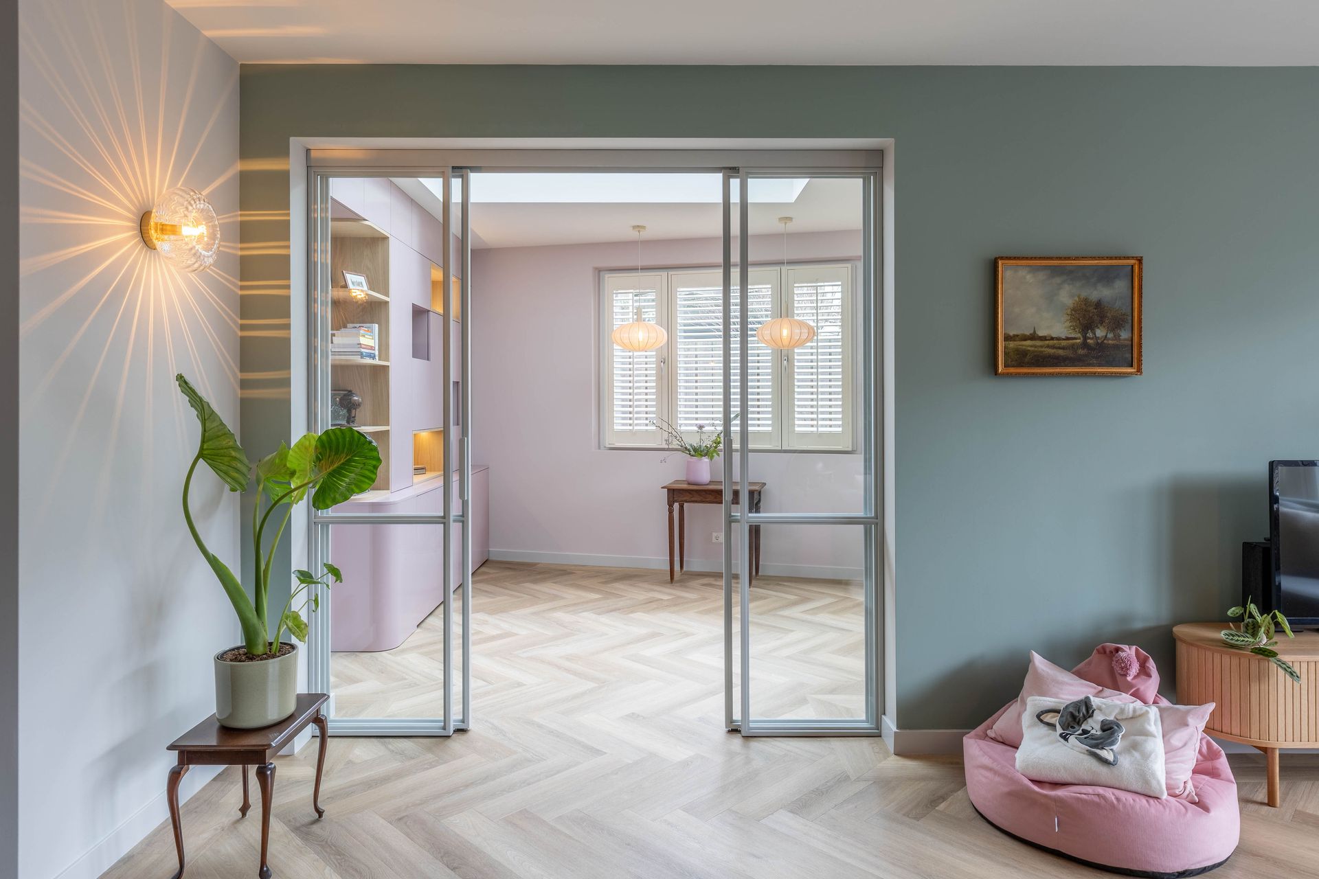 Moderne woonkamer met roze hondenmand, potplant, glazen deuren en uitzicht op een lichte keuken.