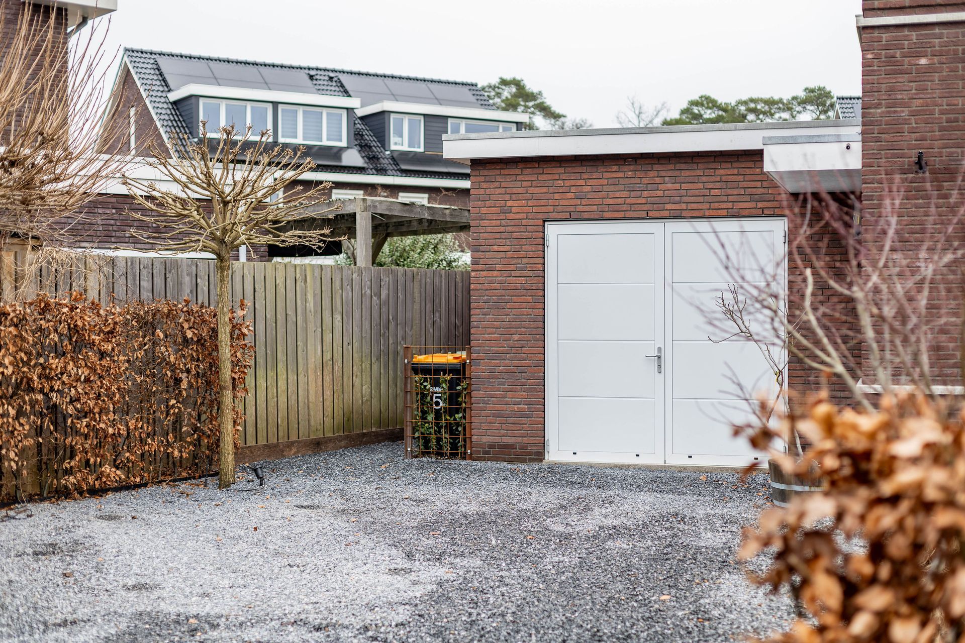 Een grindpad naast een garage van rode baksteen en een houten schutting, met huizen op de achtergrond.